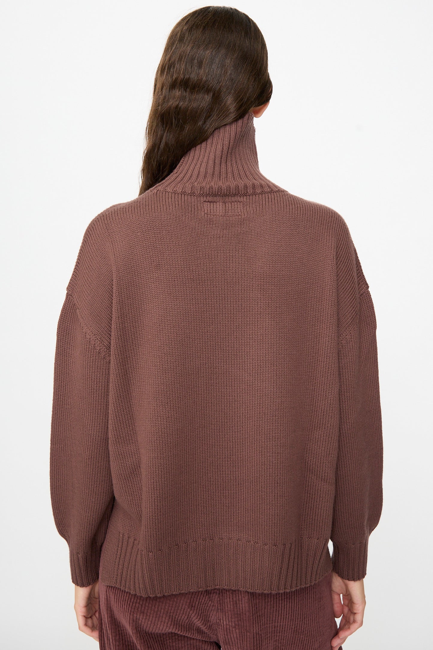 A person with long brown hair is photographed from the back against a plain white background, wearing the Labo.Art Fiore Sweater in Sasso—a loose, cashwool knitwear turtleneck.