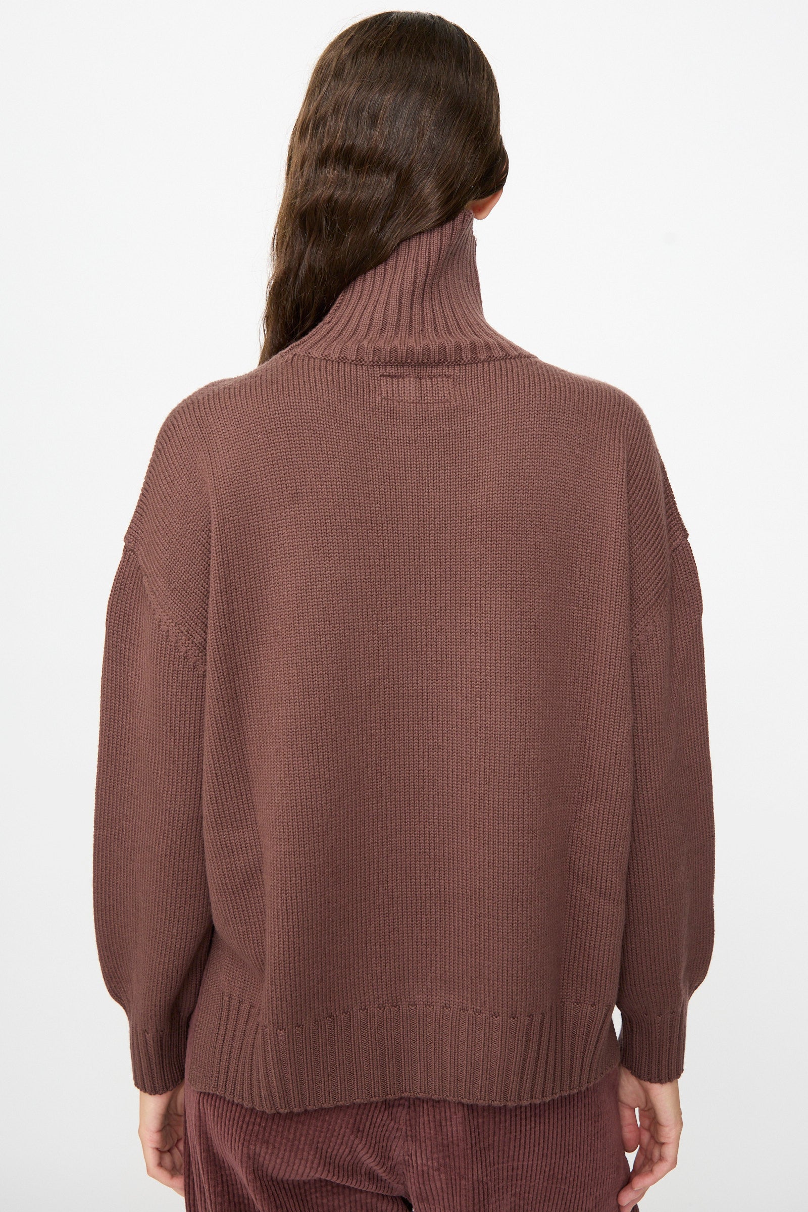 A person with long brown hair is photographed from the back against a plain white background, wearing the Labo.Art Fiore Sweater in Sasso—a loose, cashwool knitwear turtleneck.