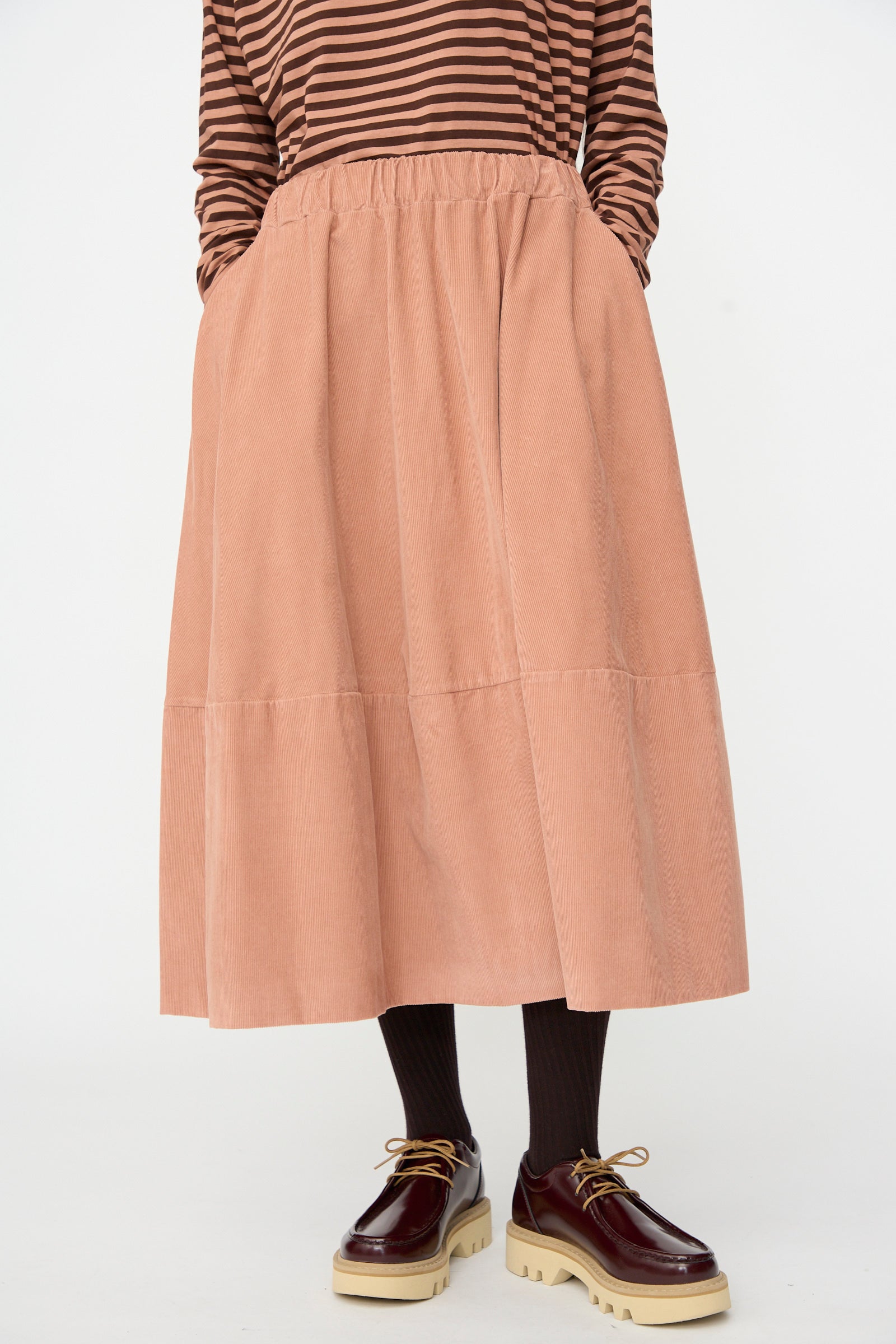 A person wearing a brown striped top, the Gea Skirt in Canyon by Labo.Art—a long peach-colored cotton midi skirt with pockets—paired with black tights and burgundy platform shoes.