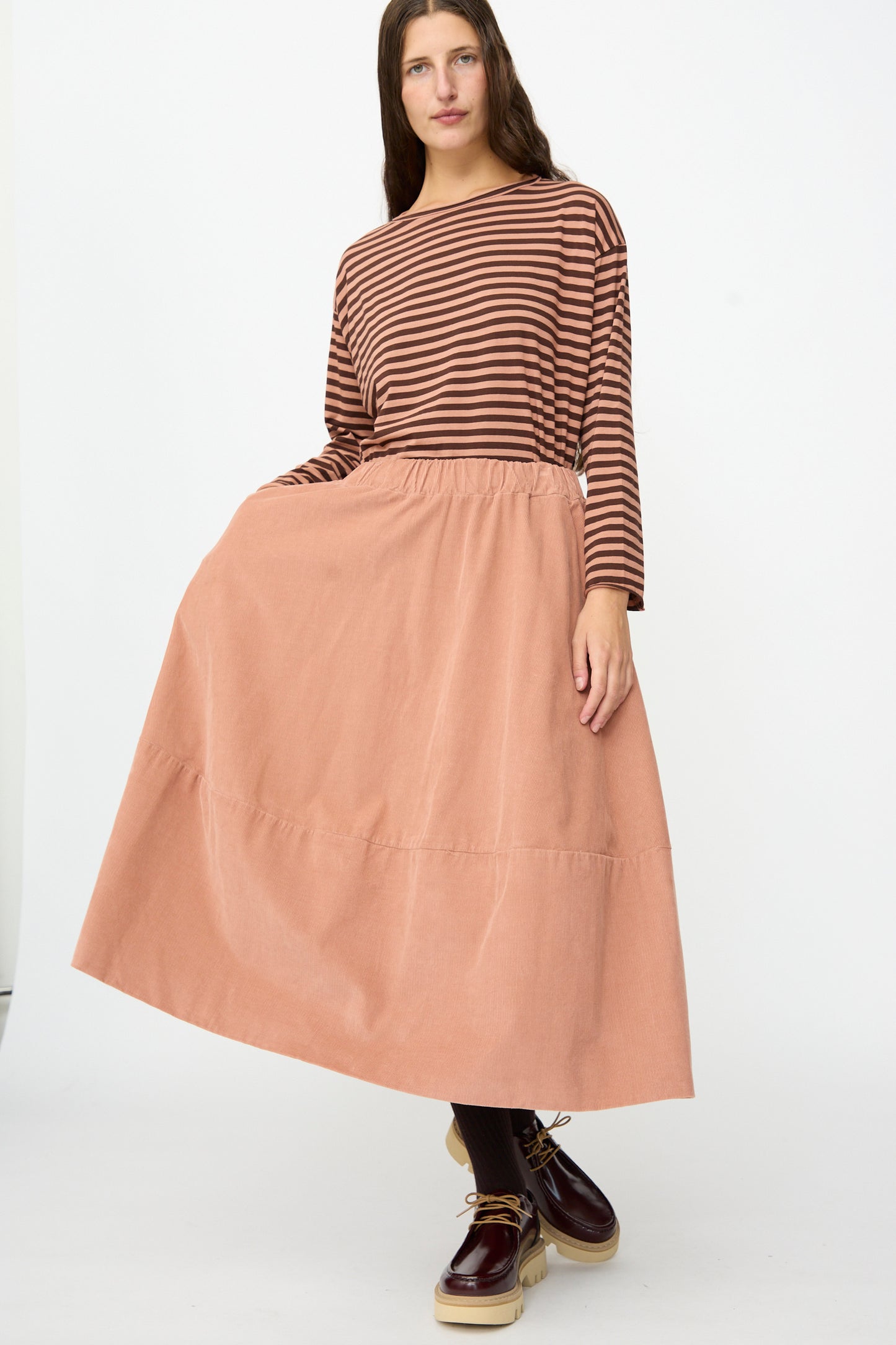 A woman stands against a plain white background wearing a brown striped long-sleeve top, dark tights, beige platform shoes, and the Labo.Art Gea Skirt in Canyon.