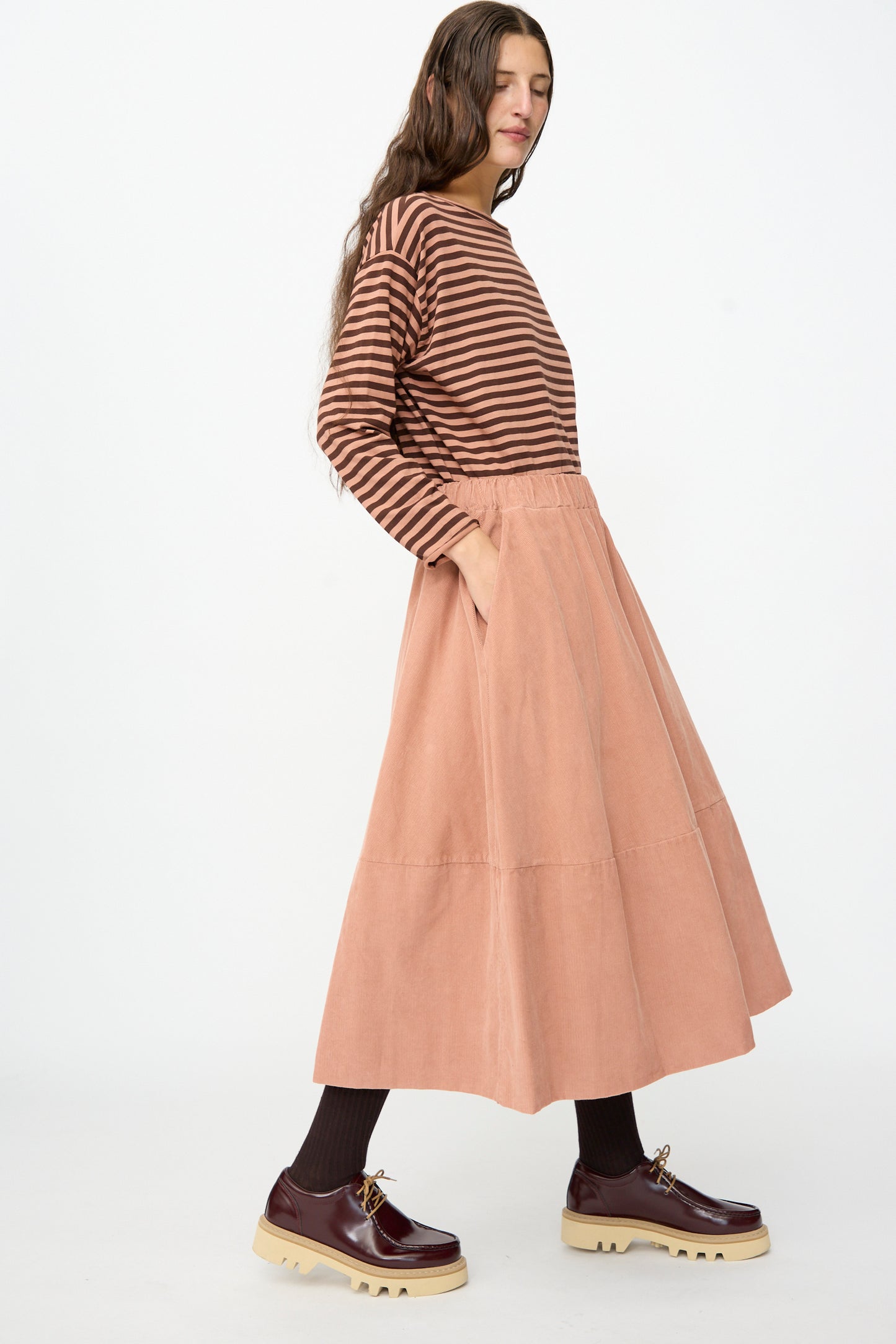 A woman in a brown striped long-sleeve top, brown socks, and platform shoes wears the Gea Skirt in Canyon by Labo.Art, standing against a plain white background.