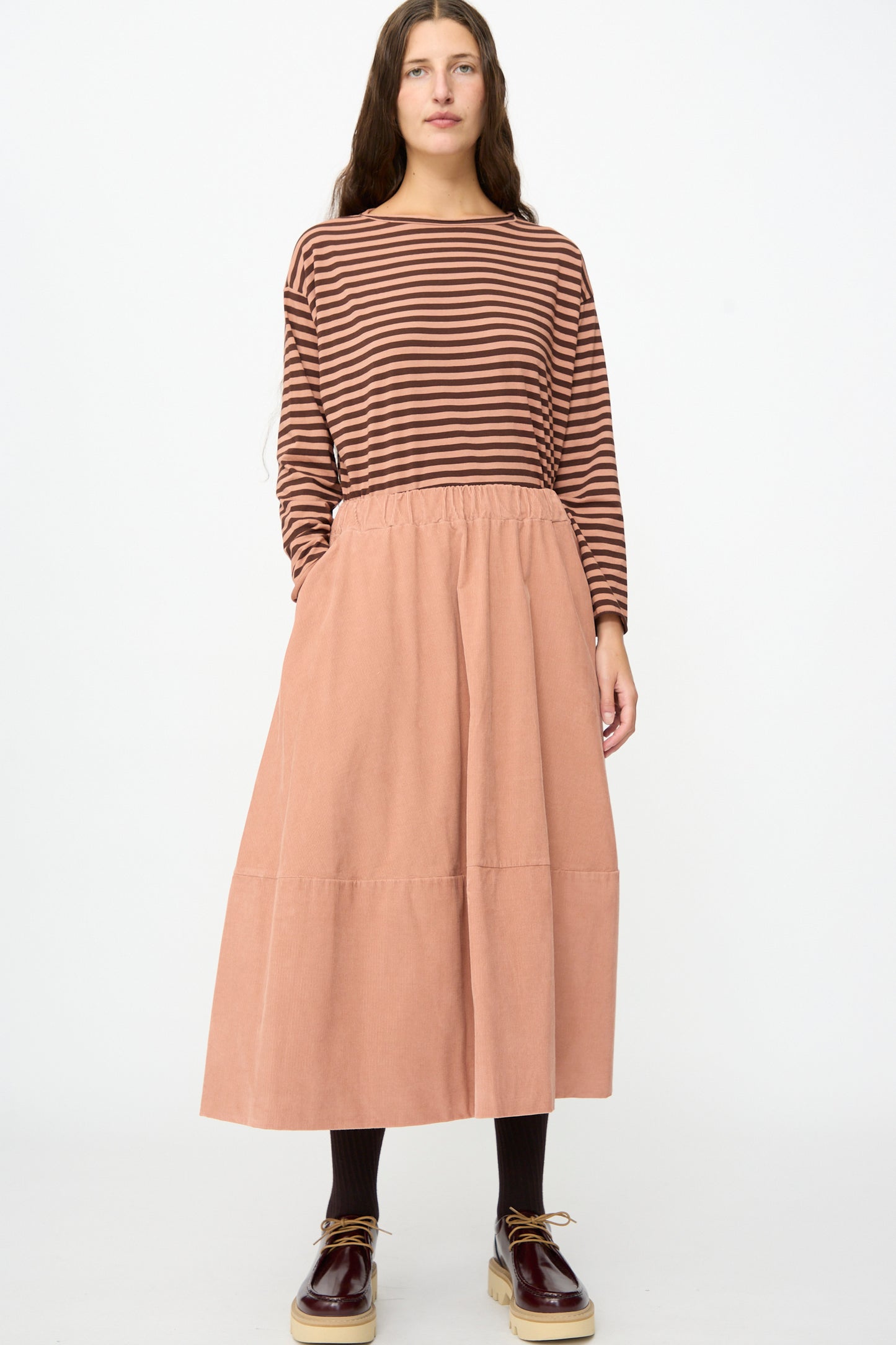A person wearing a brown striped long-sleeve top, black tights, brown shoes, and the Labo.Art Gea Skirt in Canyon stands facing forward against a plain white background.