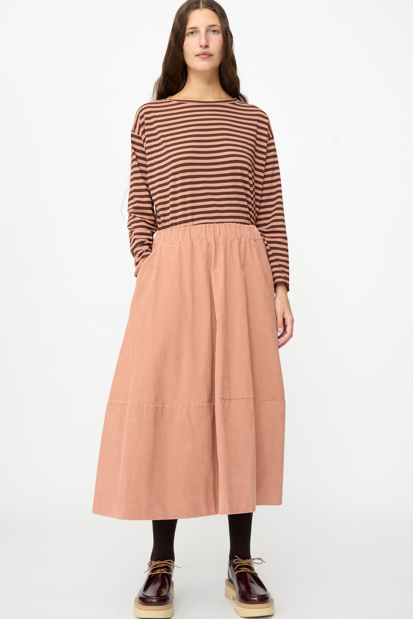 A person wearing a brown striped long-sleeve top, black tights, brown shoes, and the Labo.Art Gea Skirt in Canyon stands facing forward against a plain white background.