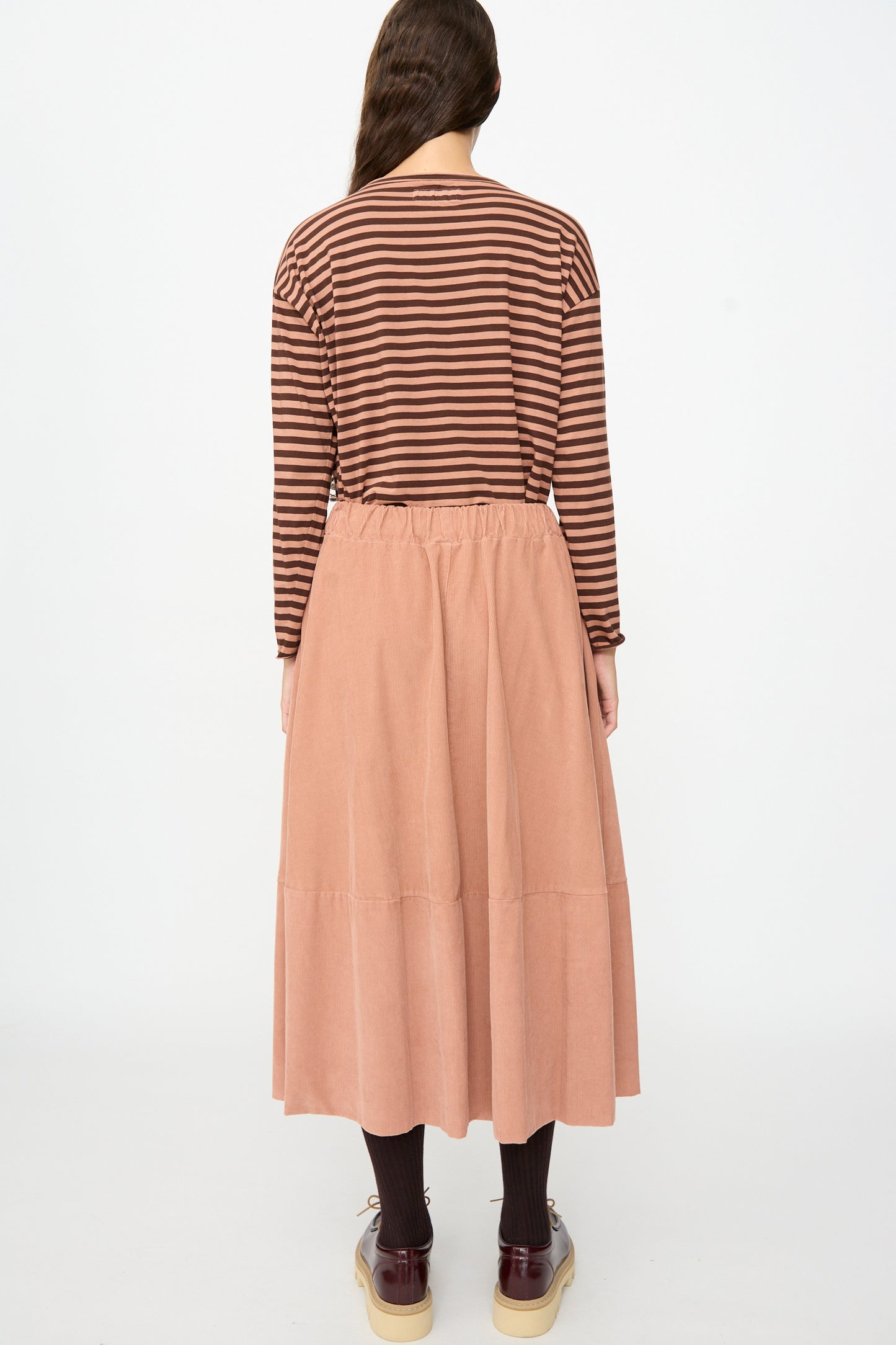 A person stands facing away, wearing a brown striped long-sleeve shirt, dark tights, brown shoes, and the Gea Skirt in Canyon by Labo.Art—a peach-colored cotton midi skirt—against a plain white background.