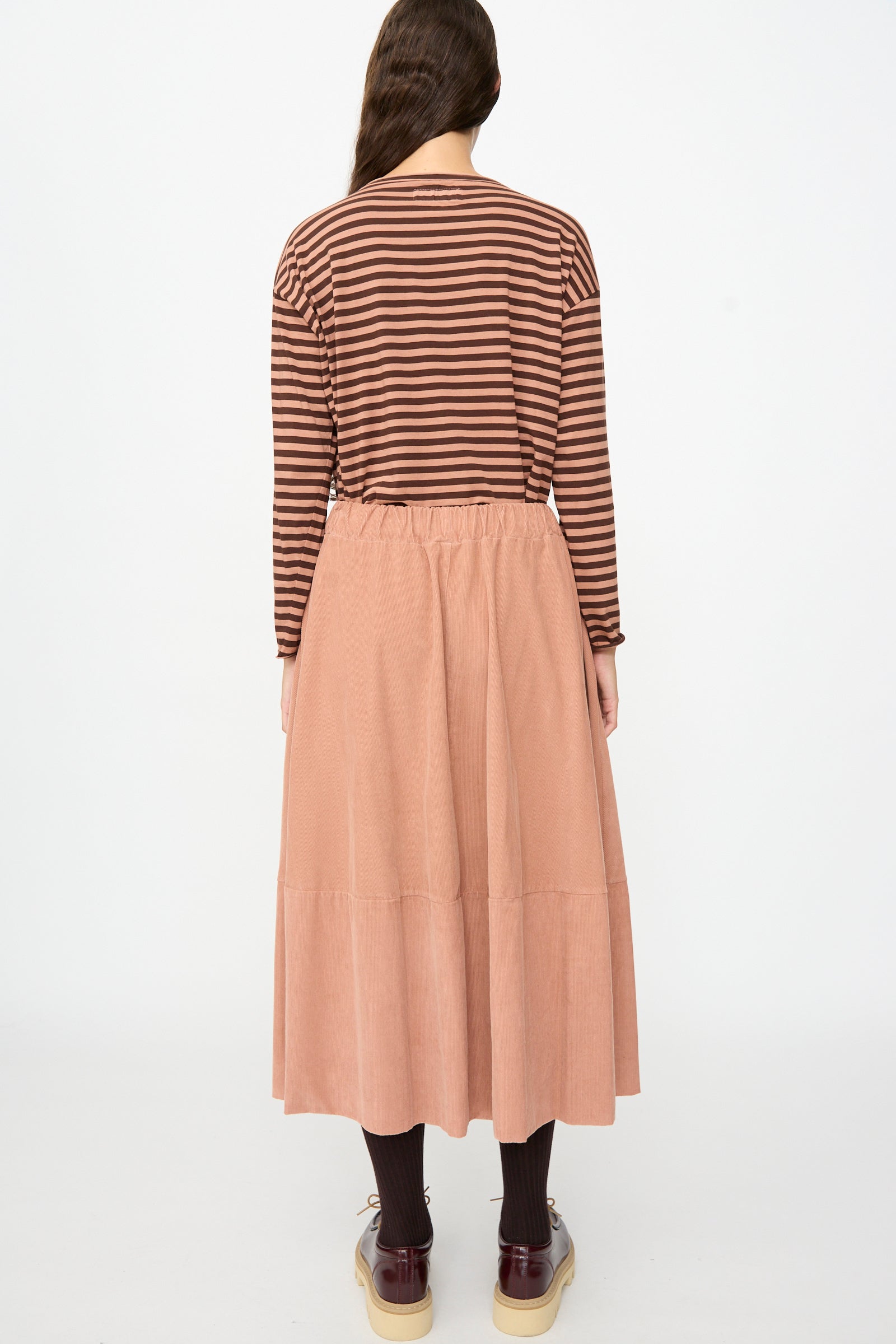 A person stands facing away, wearing a brown striped long-sleeve shirt, dark tights, brown shoes, and the Gea Skirt in Canyon by Labo.Art—a peach-colored cotton midi skirt—against a plain white background.