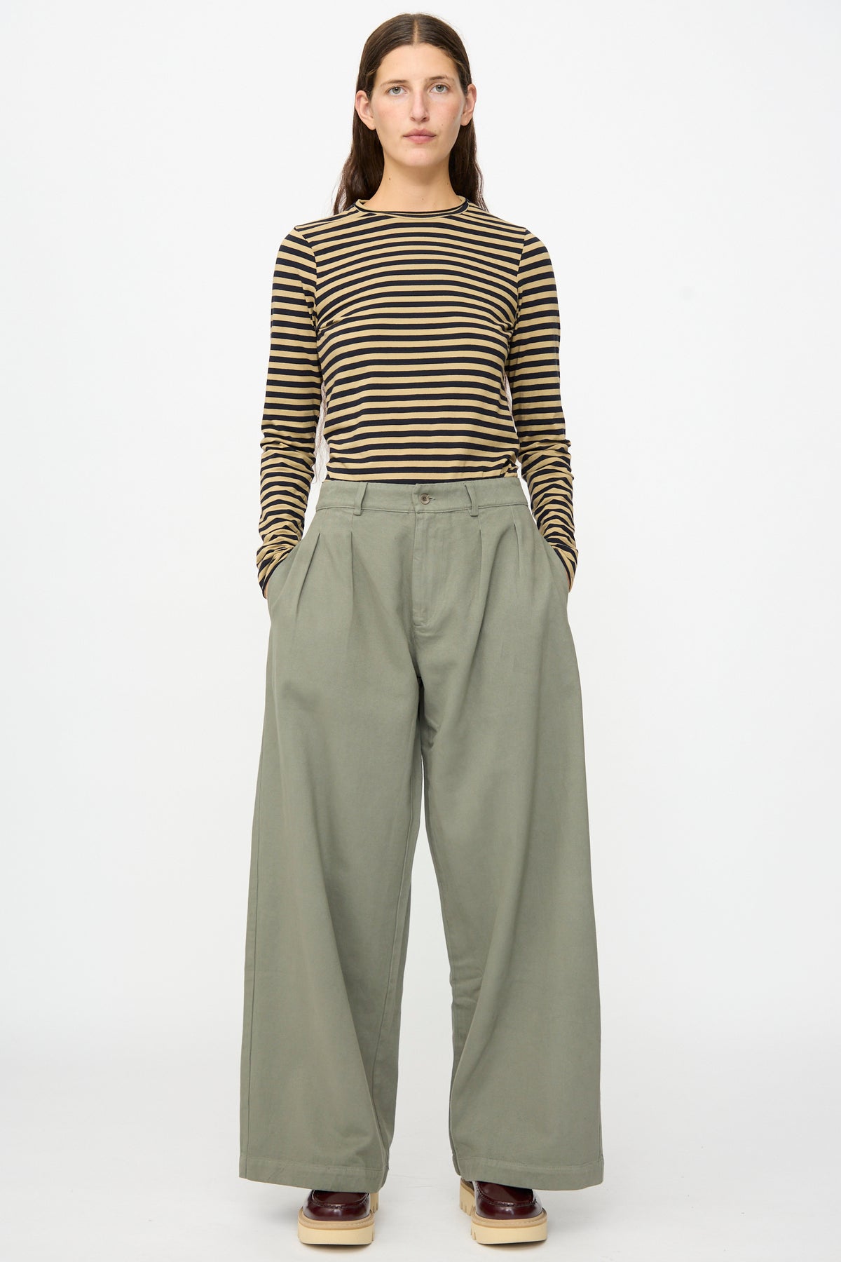 A person stands facing forward with hands in their pockets against a plain white background, wearing a black and brown striped long sleeve shirt, brown shoes, and Labo.Art’s Marlon Trouser in Lichene—wide-leg olive green brushed cotton twill pants made in Italy.