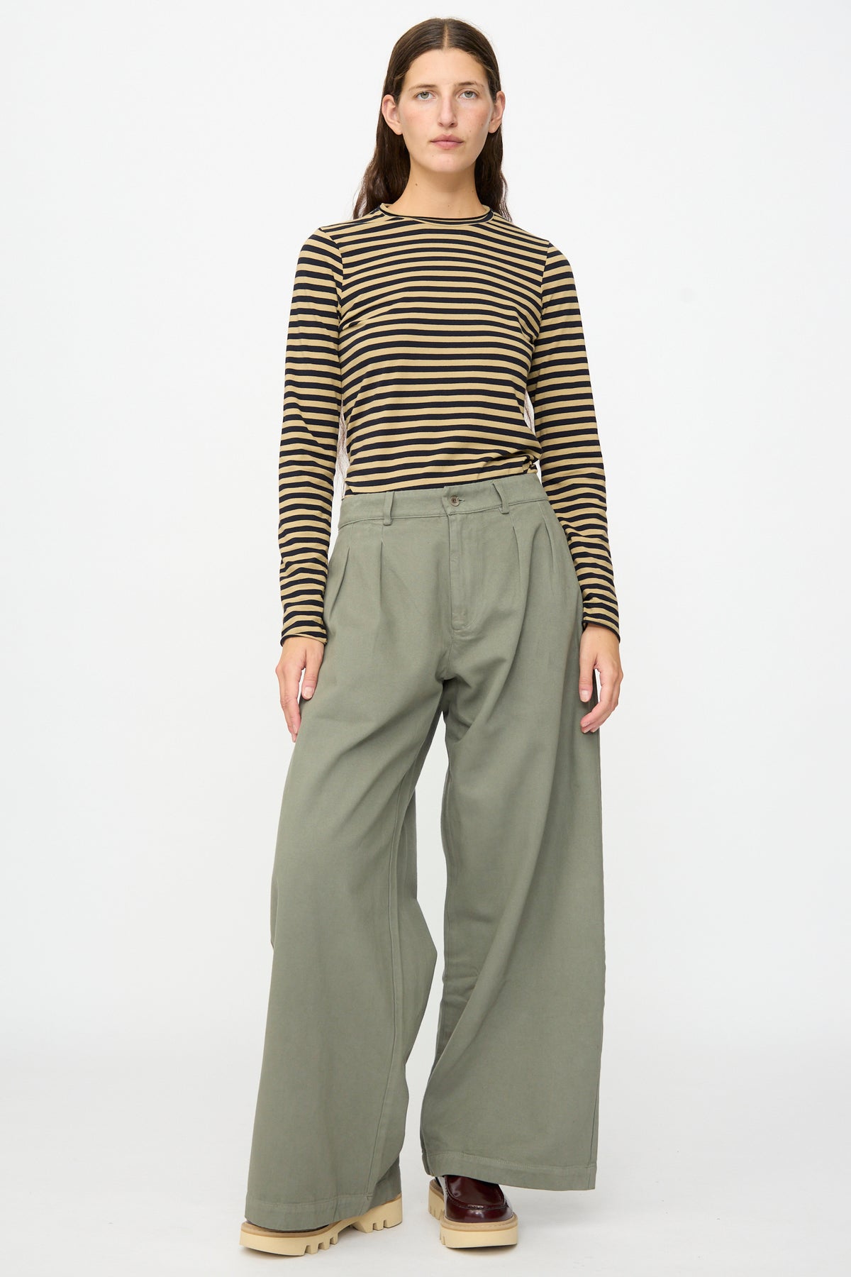 A person stands against a plain white background, wearing a long-sleeve black and yellow striped shirt, the Labo.Art Marlon Trouser in Lichene—crafted from loose olive green brushed cotton twill and made in Italy—and beige platform shoes.