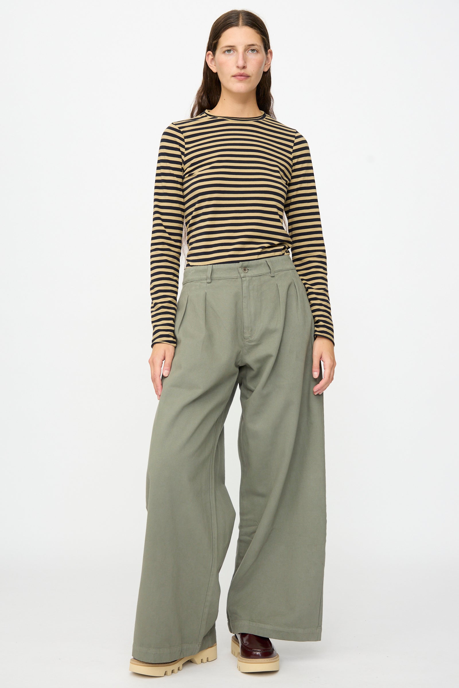 A person stands against a plain white background, wearing a long-sleeve black and yellow striped shirt, the Labo.Art Marlon Trouser in Lichene—crafted from loose olive green brushed cotton twill and made in Italy—and beige platform shoes.