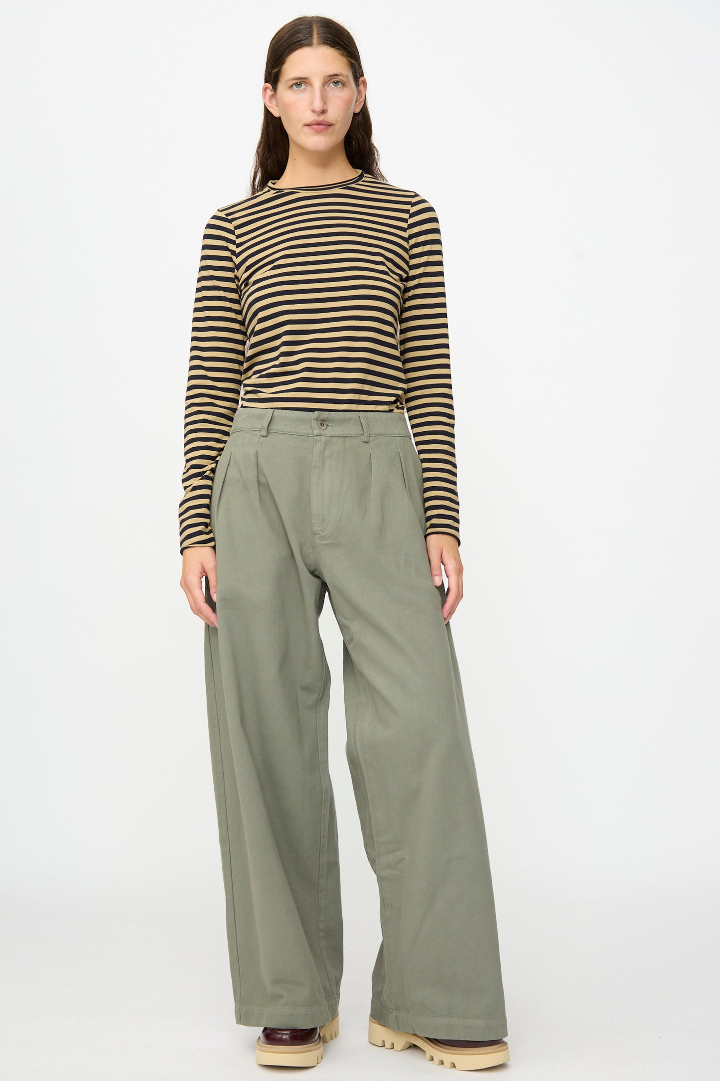 A person stands facing forward against a plain white background, wearing a brown and black striped long-sleeve shirt, Labo.Art Marlon Trouser in Lichene featuring a sage green brushed cotton twill with a wide-leg silhouette, and beige platform shoes.