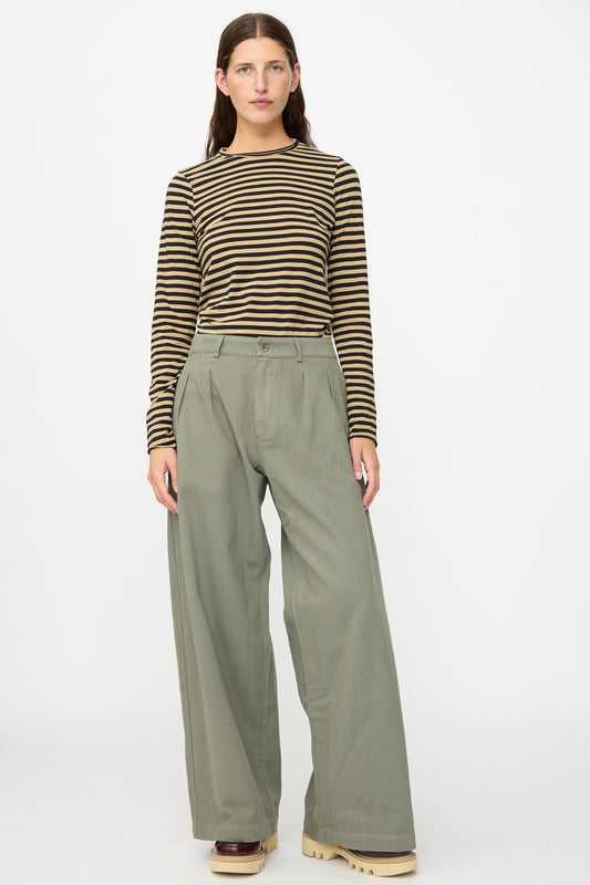 A person stands facing forward against a plain white background, wearing a brown and black striped long-sleeve shirt, Labo.Art Marlon Trouser in Lichene featuring a sage green brushed cotton twill with a wide-leg silhouette, and beige platform shoes.