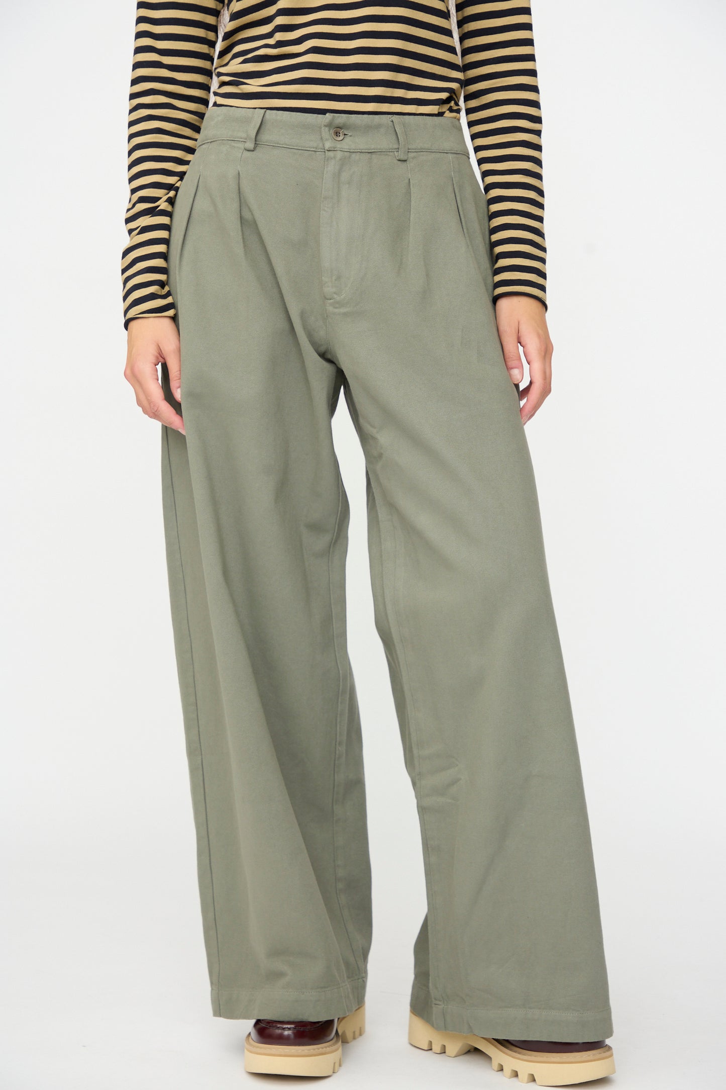 A person wearing the Labo.Art Marlon Trouser in Lichene—olive green wide-leg trousers crafted from brushed cotton twill—paired with a brown and black striped long-sleeve top and chunky beige-soled shoes, standing against a plain white background.