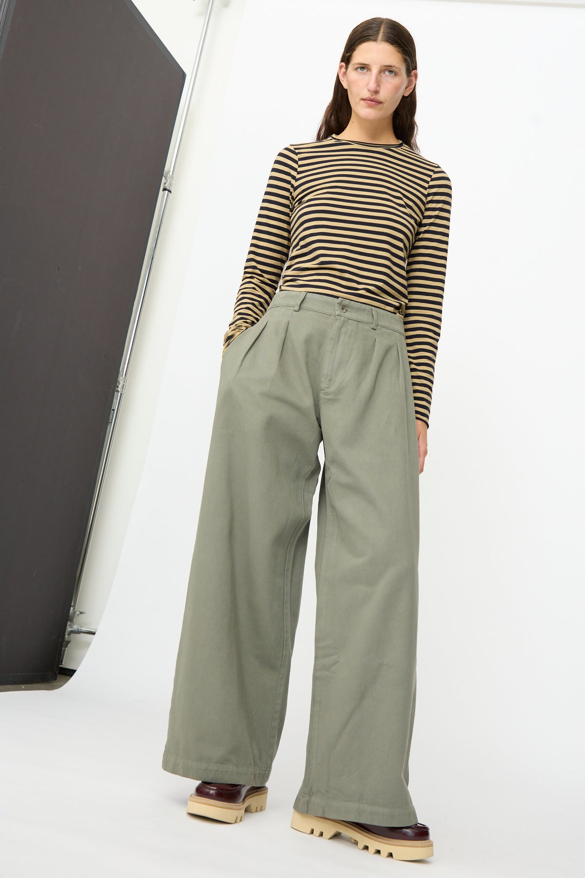 A person stands against a white background, wearing a brown and black striped long-sleeve shirt, the Marlon Trouser in Lichene by Labo.Art—loose, high-waisted brushed cotton twill pants in olive green—and beige platform shoes.
