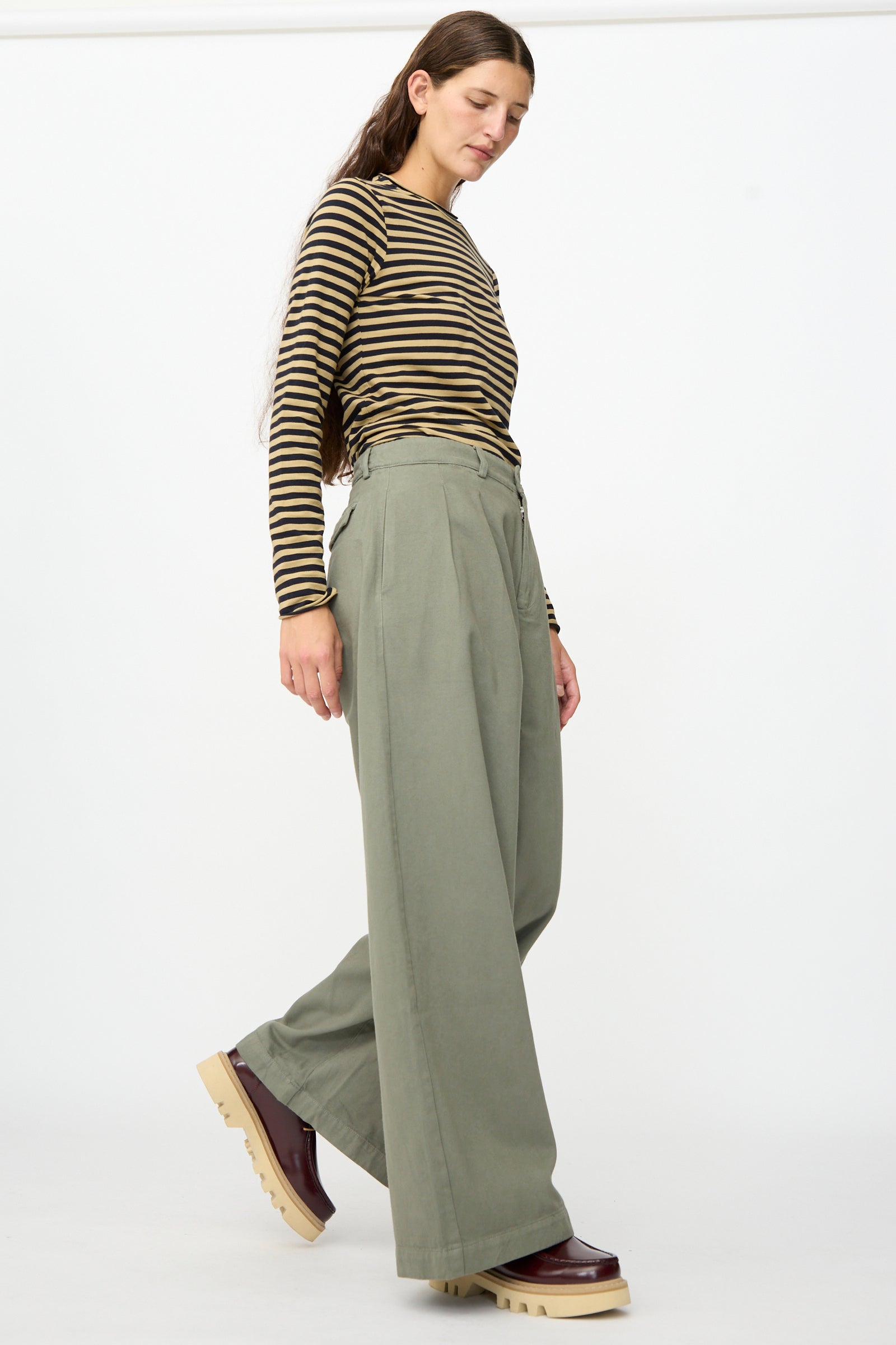 A person stands against a plain white background wearing a brown striped long-sleeve shirt, Labo.Art’s Marlon Trouser in Lichene featuring a brushed cotton twill fabric and wide-leg silhouette, paired with brown platform shoes.