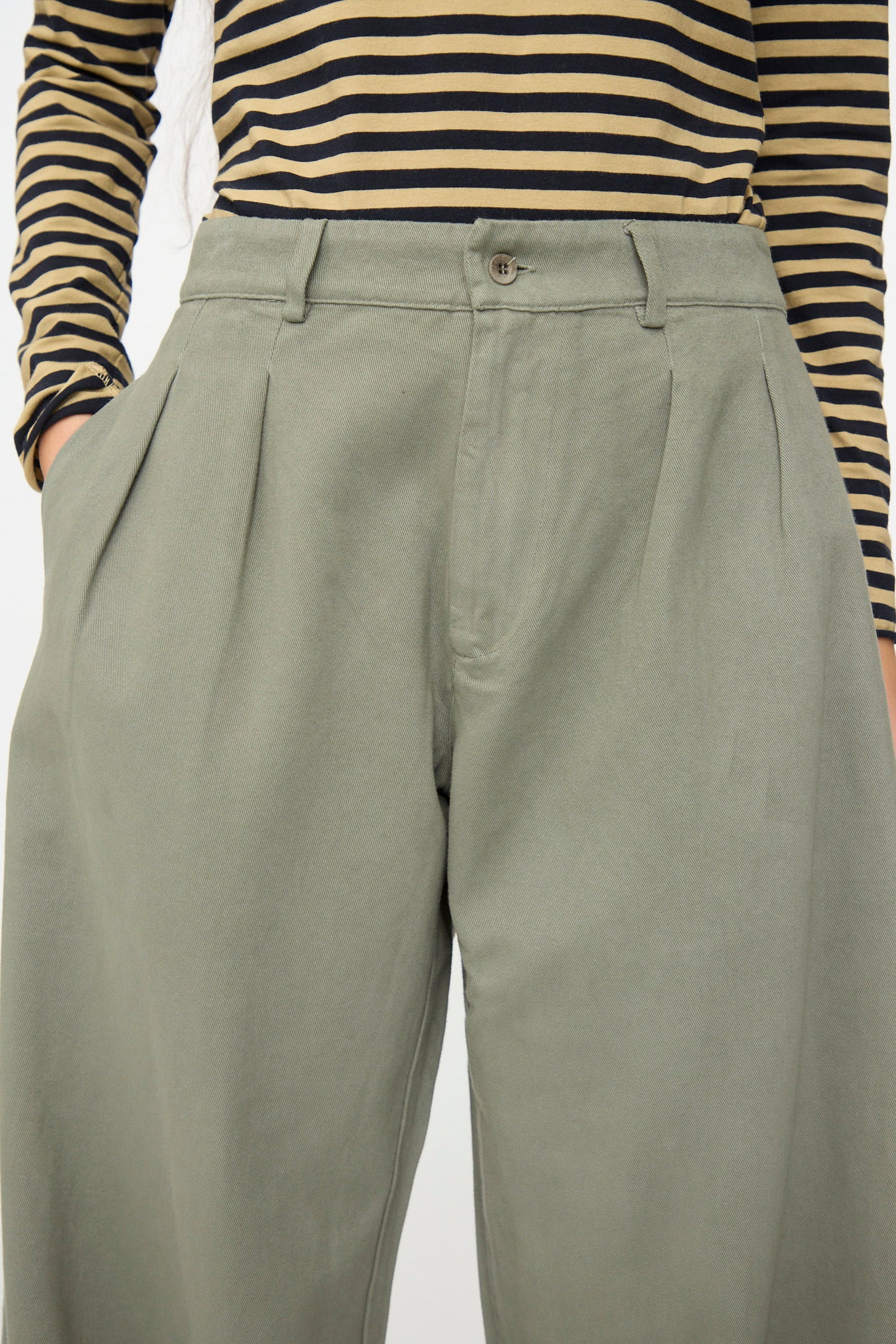 Person wearing a black and tan striped long-sleeve shirt with high-waisted, pleated Labo.Art Marlon Trouser in Lichene, hands in pockets.