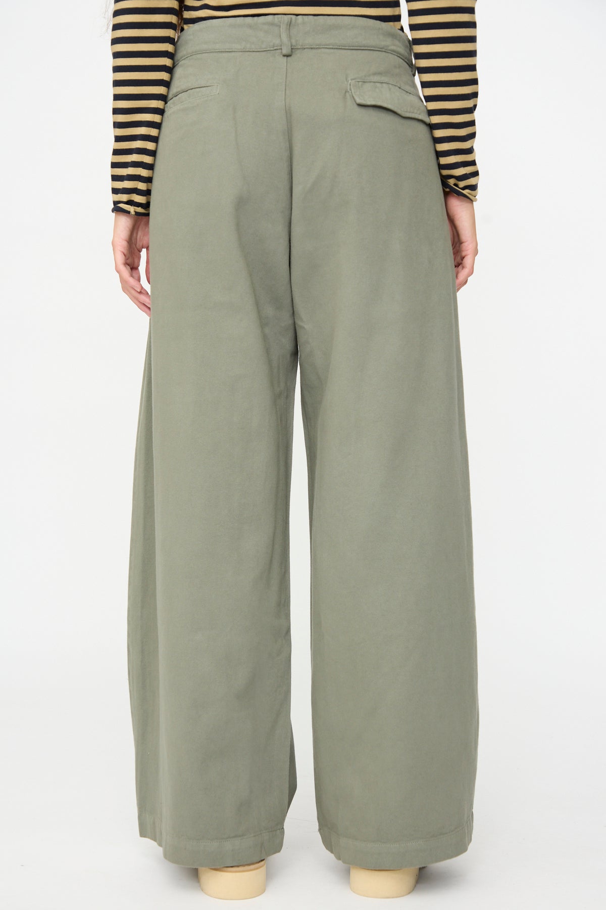 A person seen from the back against a plain white background wears the Labo.Art Marlon Trouser in Lichene—a high-waisted, wide-leg olive green brushed cotton twill pant—paired with a black and tan striped long-sleeve top.