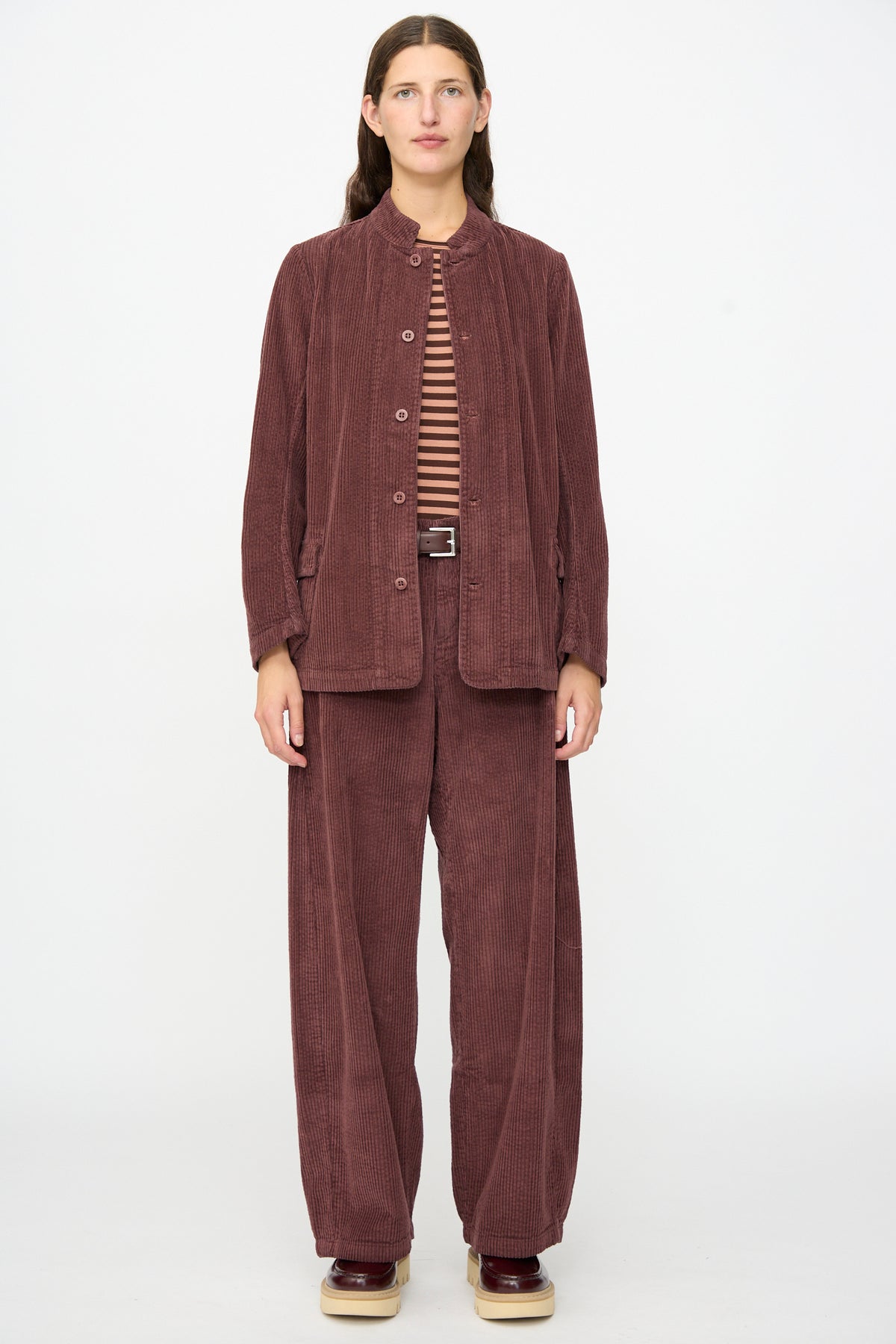 A person stands against a white background, wearing the Labo.Art Pina Jacket in Sasso—a relaxed maroon cotton corduroy jacket—paired with matching wide-leg pants, a striped shirt, belt, and platform shoes. This expertly crafted piece offers both comfort and style.