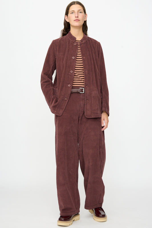 A person stands against a plain white background wearing the Labo.Art Pina Jacket in Sasso, a loose-fitting maroon cotton corduroy jacket made in Italy, styled as part of a suit layered over a brown striped top and paired with beige platform shoes.