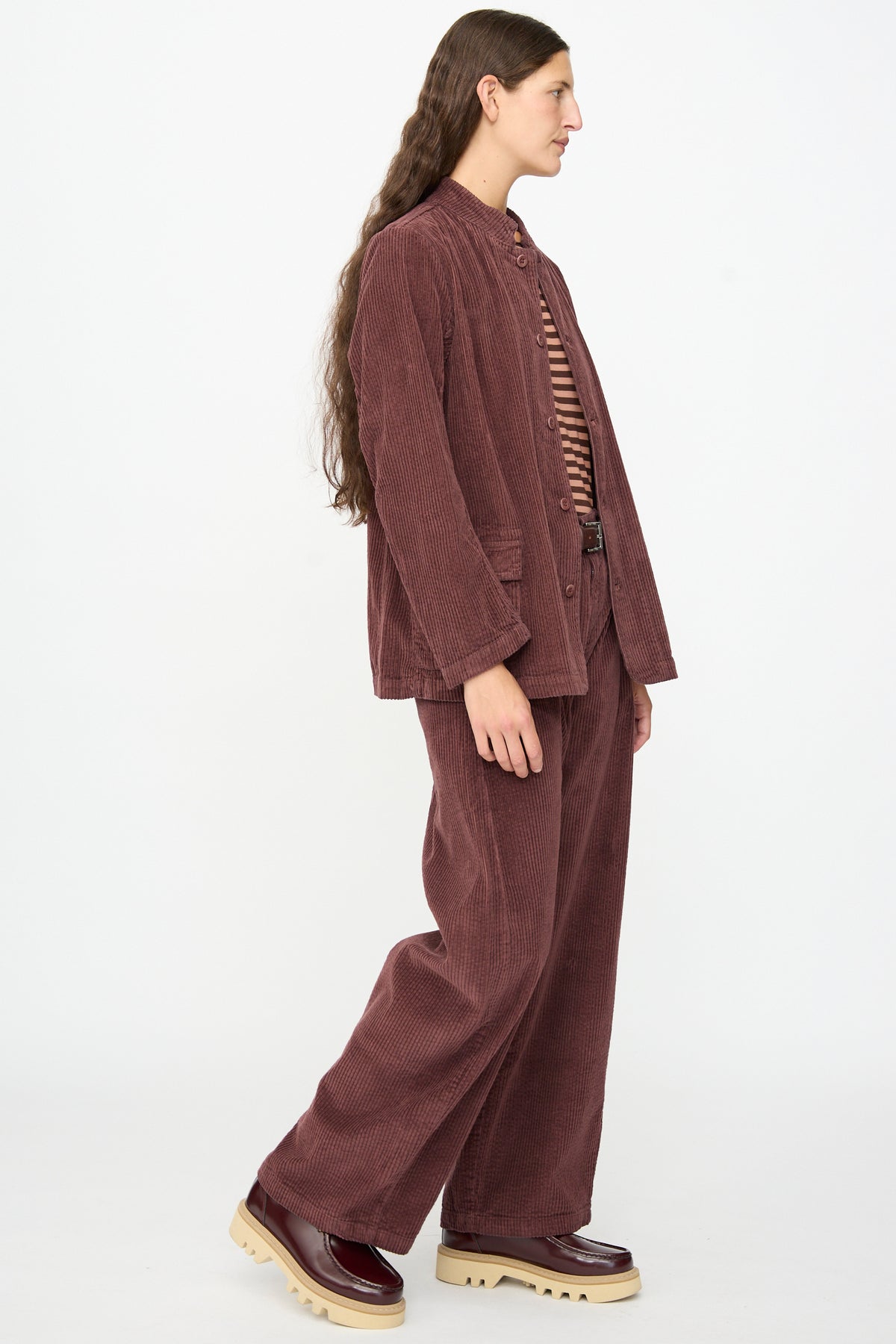 A person with long hair wears the Pina Jacket in Sasso by Labo.Art—crafted from burgundy cotton corduroy in Italy—styled with a striped shirt and chunky sole shoes, standing in profile against a plain white background.