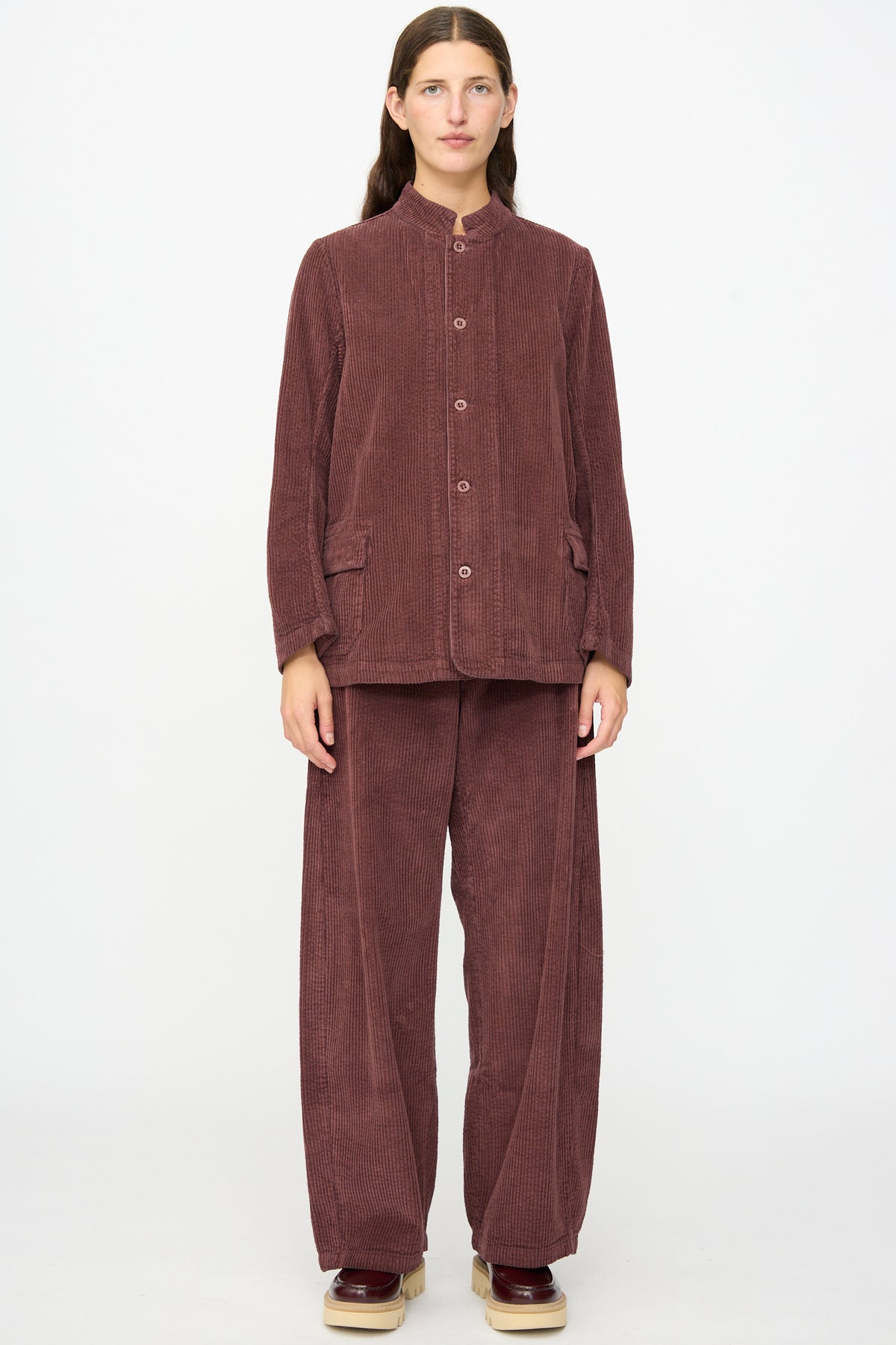 A person stands against a plain background wearing the Labo.Art Pina Jacket in Sasso, a loose-fitting mauve cotton corduroy piece with a buttoned front, styled with wide-leg pants and platform shoes. The relaxed silhouette of the jacket adds effortless style to the ensemble.