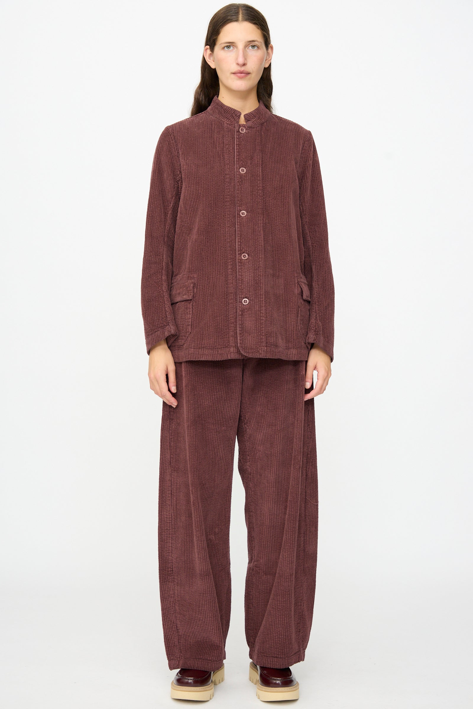 A person stands against a plain background wearing the Labo.Art Pina Jacket in Sasso, a loose-fitting mauve cotton corduroy piece with a buttoned front, styled with wide-leg pants and platform shoes. The relaxed silhouette of the jacket adds effortless style to the ensemble.