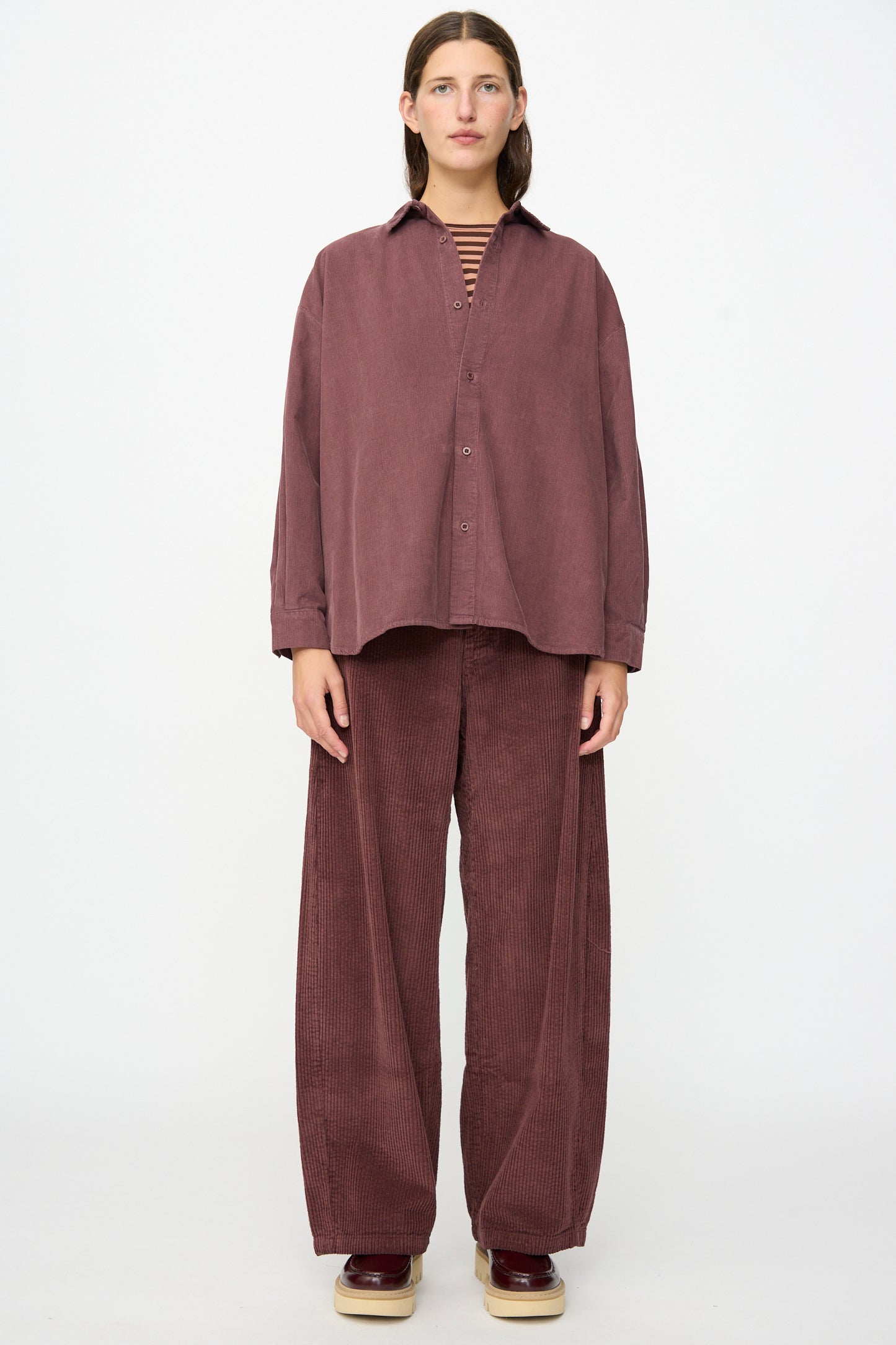 A person stands facing forward in a relaxed fit, mauve cotton needlecord Quadra Shirt in Sasso by Labo.Art and matching wide-leg pants, with a striped inner shirt and platform shoes, against a plain white background.