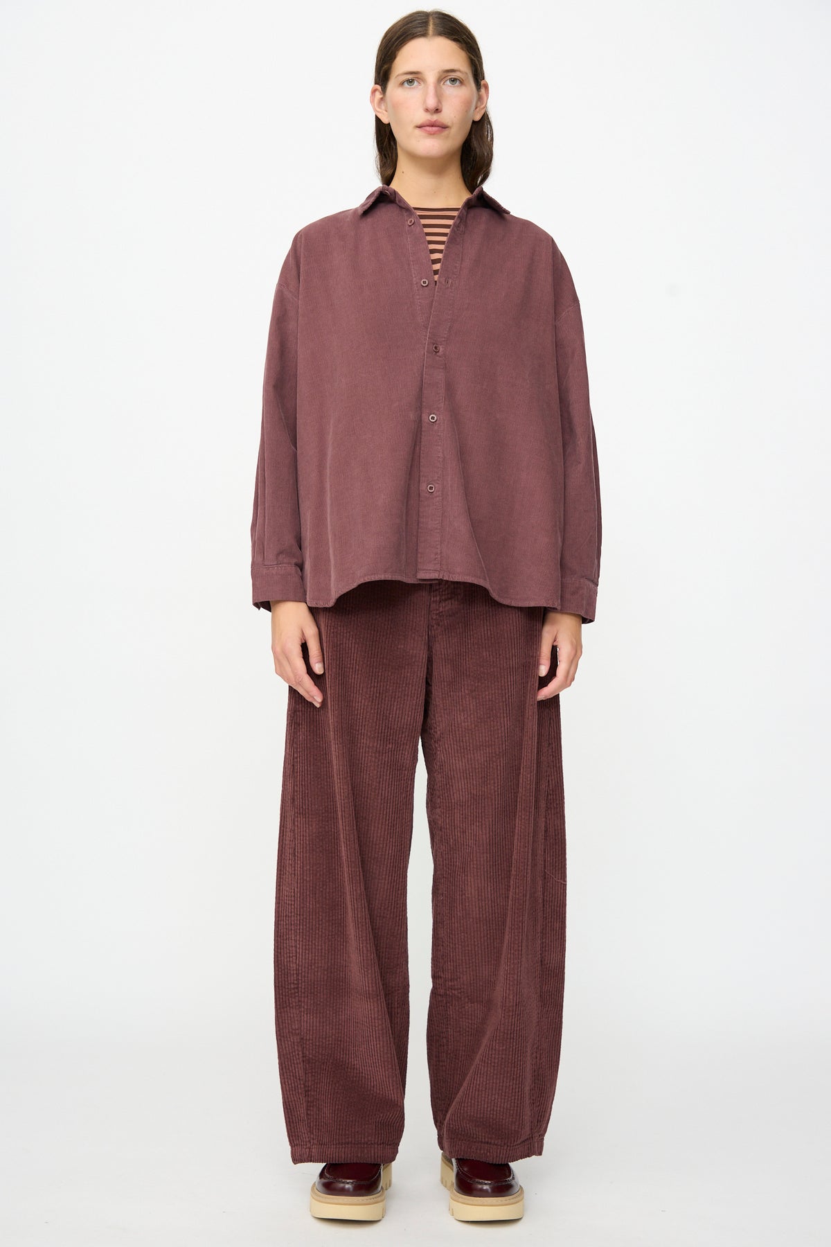 A person stands facing forward in a relaxed fit, mauve cotton needlecord Quadra Shirt in Sasso by Labo.Art and matching wide-leg pants, with a striped inner shirt and platform shoes, against a plain white background.