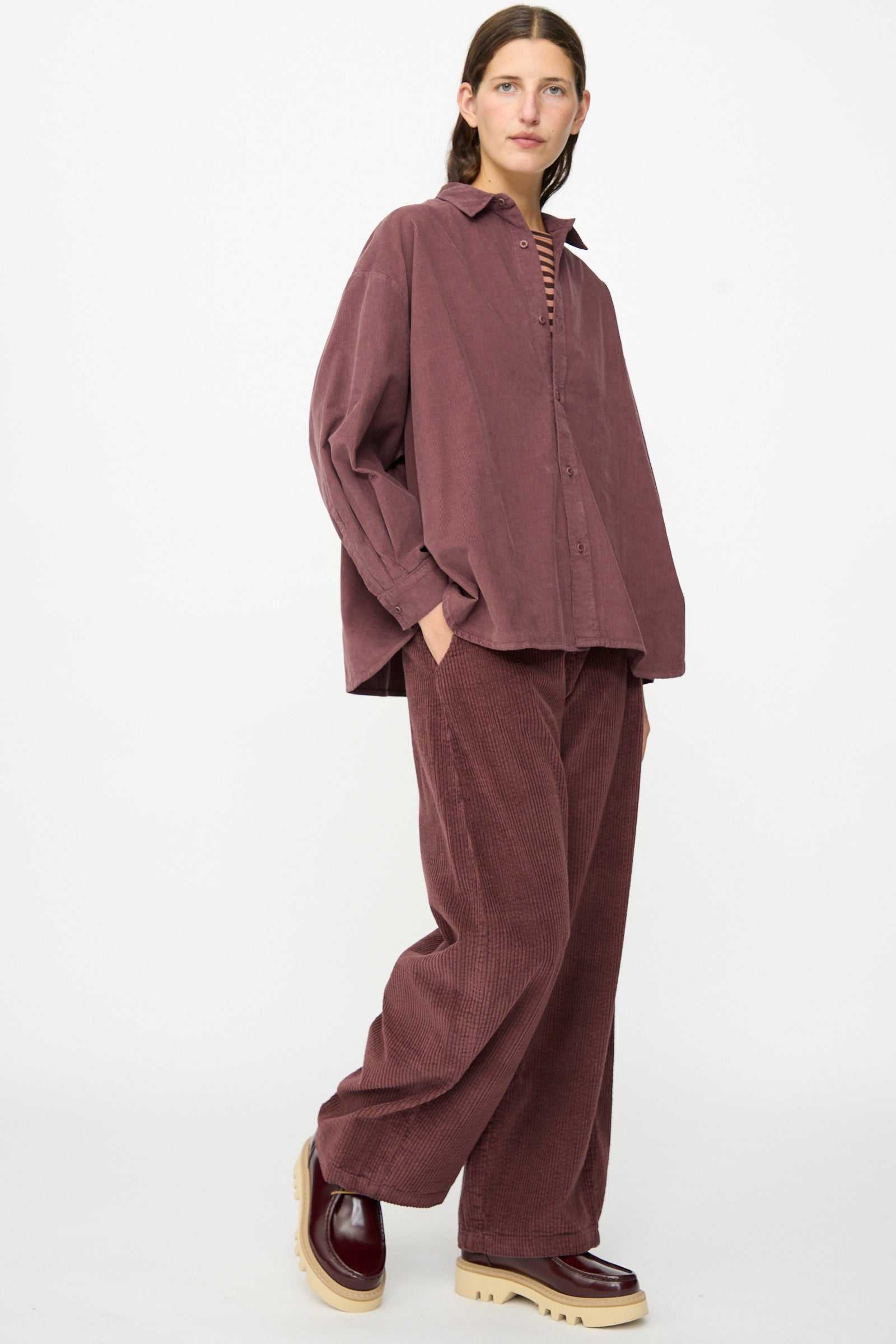 Wearing the Labo.Art Quadra Shirt in Sasso, a relaxed fit mauve cotton needlecord button-down, and matching pants, paired with brown platform shoes, the person stands against a plain white background.