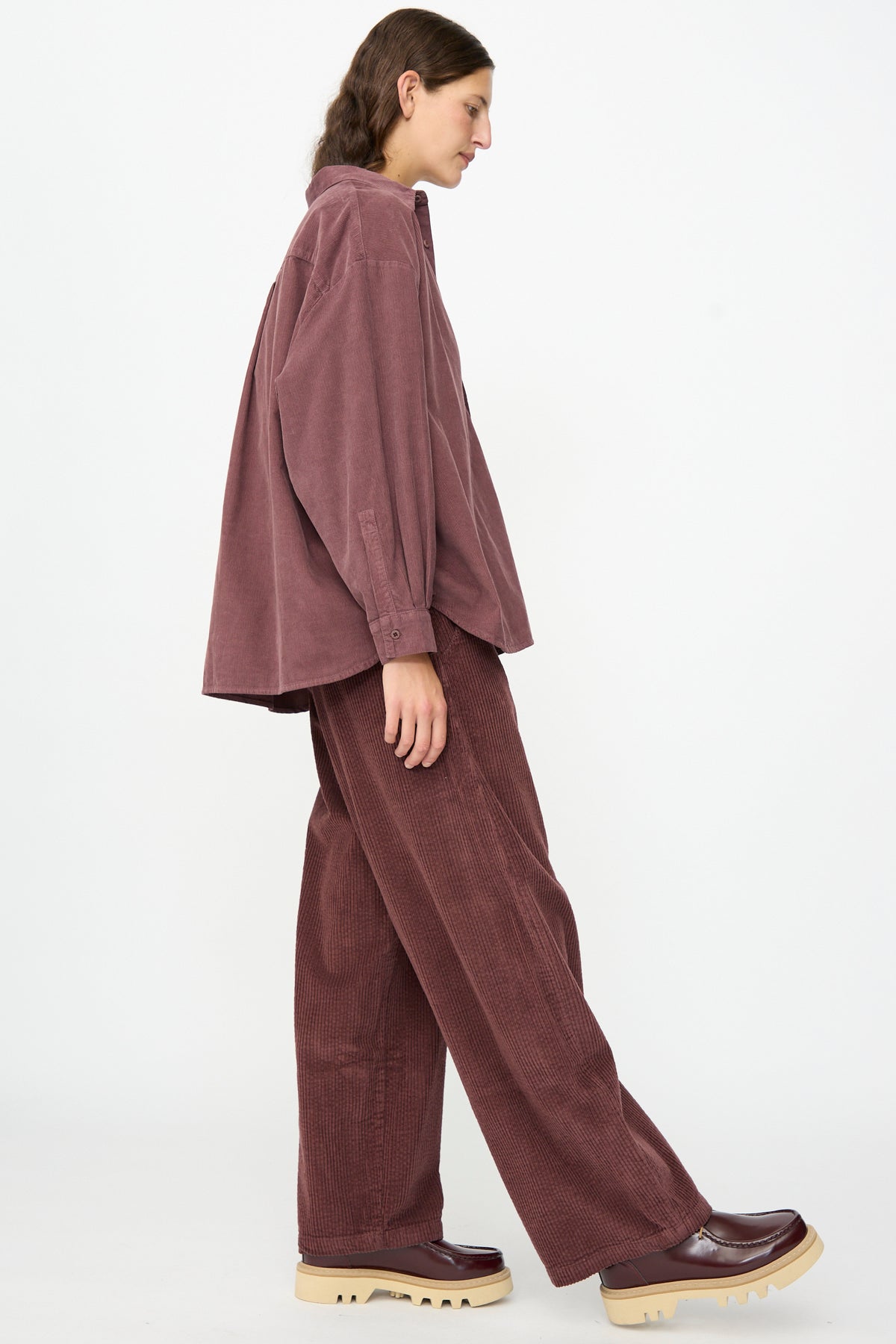 A person stands sideways against a plain white background wearing the Labo.Art Quadra Shirt in Sasso, a relaxed fit dark mauve cotton needlecord button-down shirt paired with matching pants and chunky beige-soled shoes.