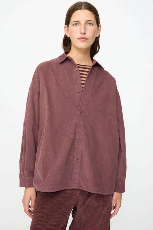 Wearing the Labo.Art Quadra Shirt in Sasso—a relaxed fit maroon cotton needlecord button-down—layered over a striped top and paired with matching maroon corduroy pants, the person stands against a plain white background.