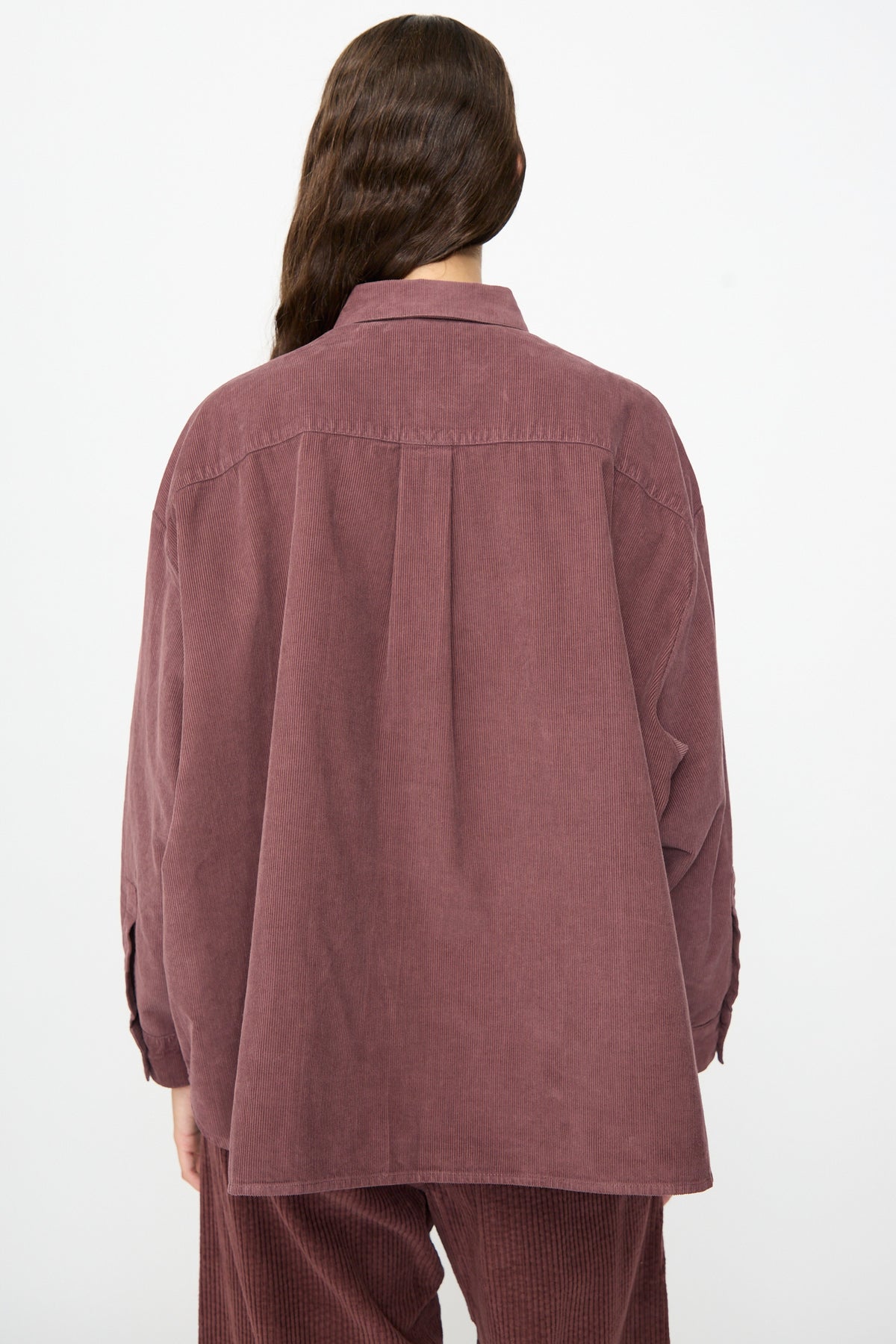 A person with long brown hair is shown from the back against a plain white background, wearing the Labo.Art Quadra Shirt in Sasso—a relaxed fit, mauve cotton needlecord button down—paired with matching pants.