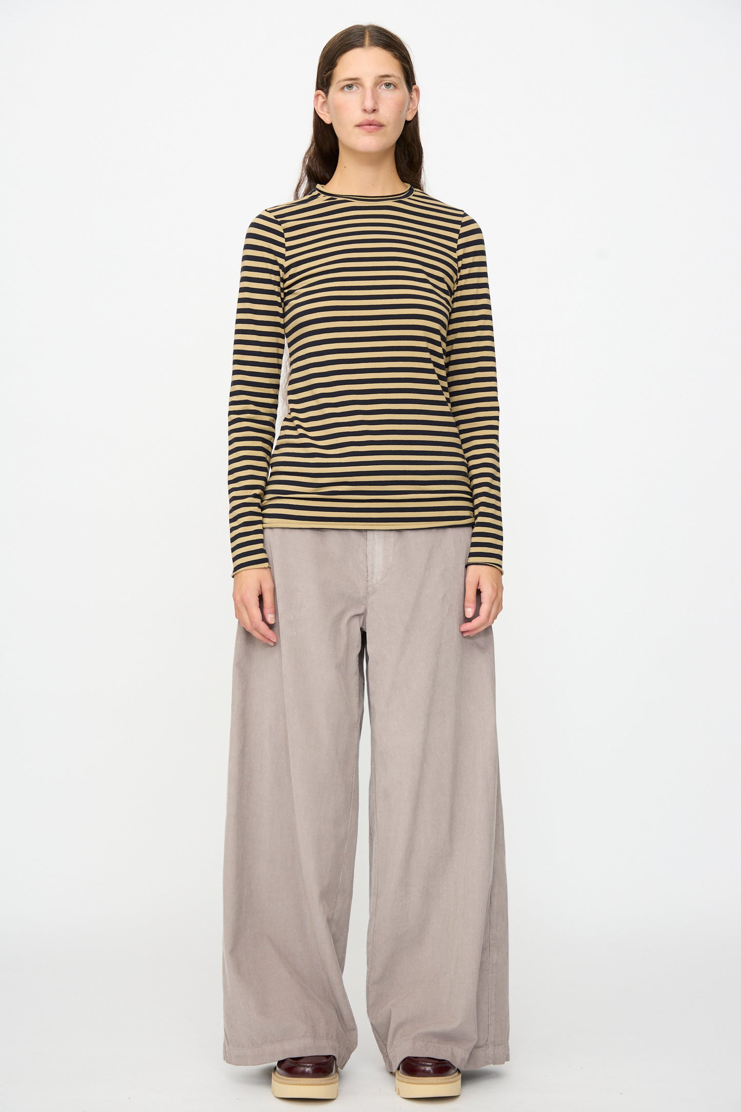 A person stands against a white background, wearing the Labo.Art Striped Jersey Basic Longsleeve in Fennec—a black and olive classic cotton crewneck—paired with wide-leg beige pants and brown shoes.