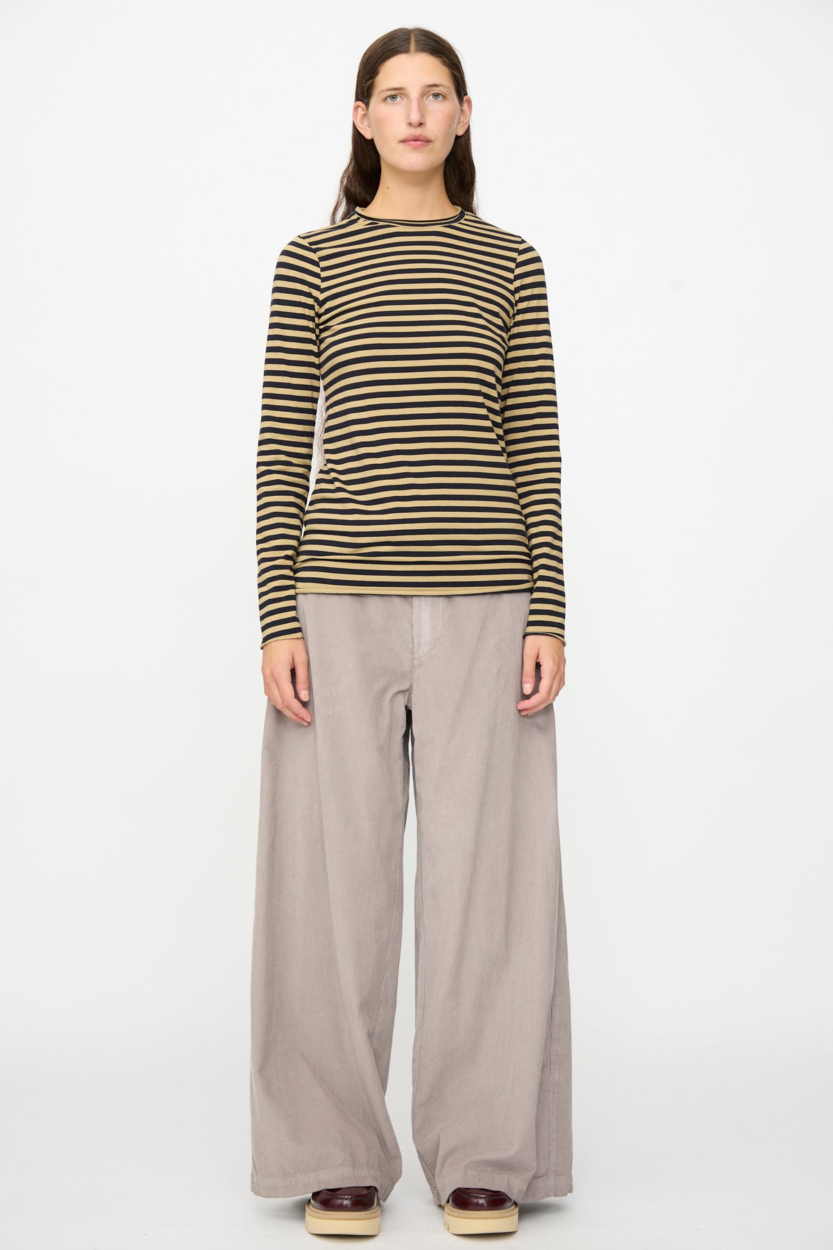 A person stands against a white background, wearing the Labo.Art Striped Jersey Basic Longsleeve in Fennec—a black and olive classic cotton crewneck—paired with wide-leg beige pants and brown shoes.
