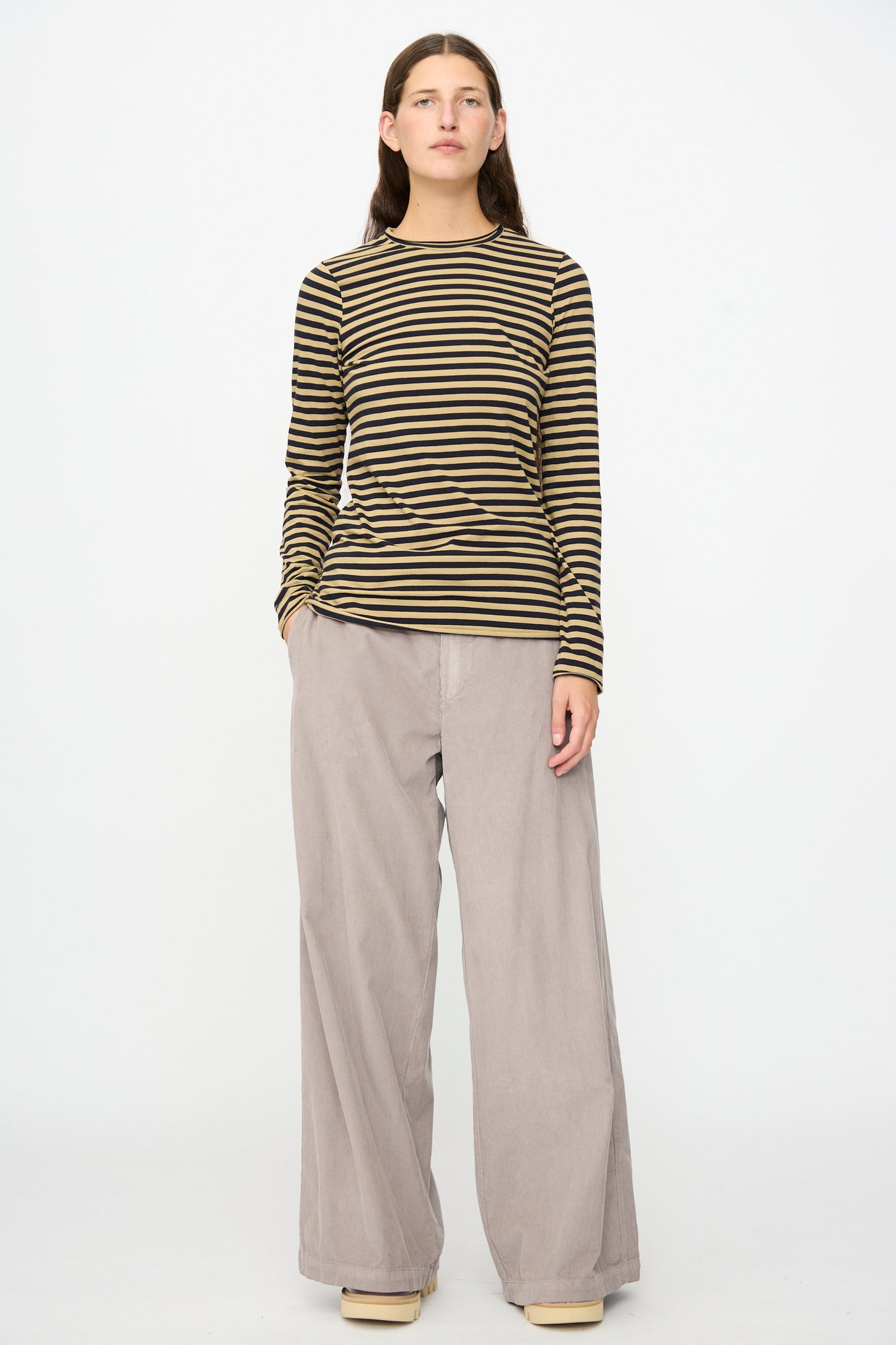 A person stands against a white background wearing the Labo.Art Striped Jersey Basic Longsleeve in Fennec paired with loose, wide-leg beige pants and light-colored platform shoes.