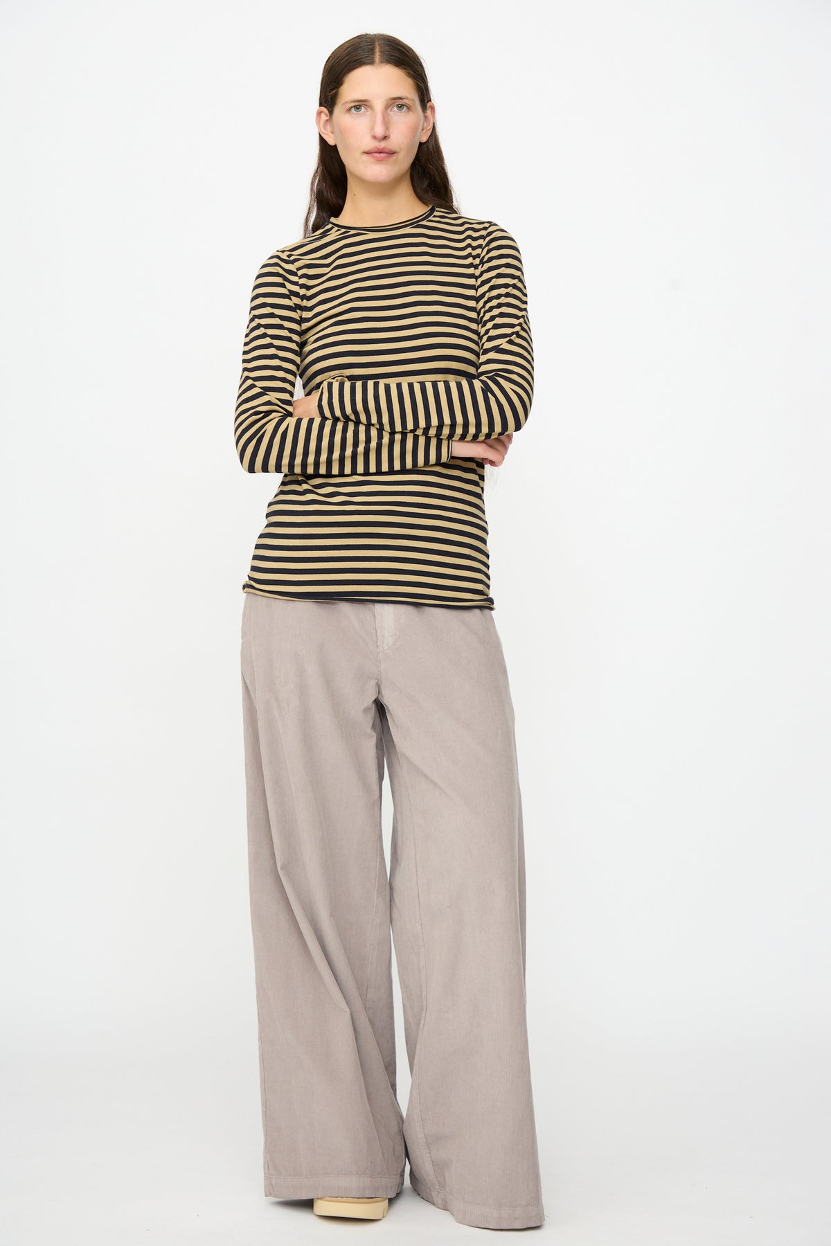 A person stands with arms crossed, wearing the Labo.Art Striped Jersey Basic Longsleeve in Fennec paired with wide-leg beige pants, set against a plain white background.