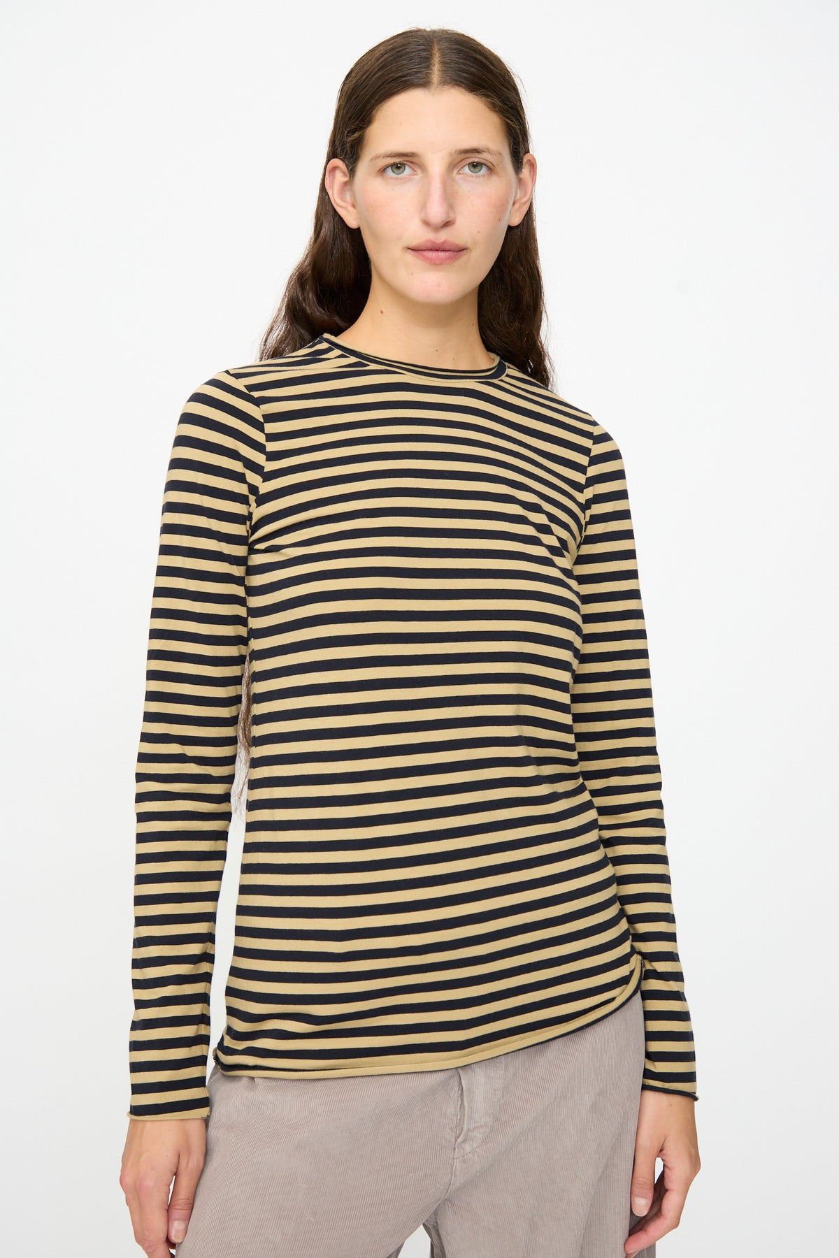 A person with long brown hair is wearing the Labo.Art Striped Jersey Basic Longsleeve in Fennec, a slim-fit beige and black striped top, paired with light beige pants, standing against a plain white background.