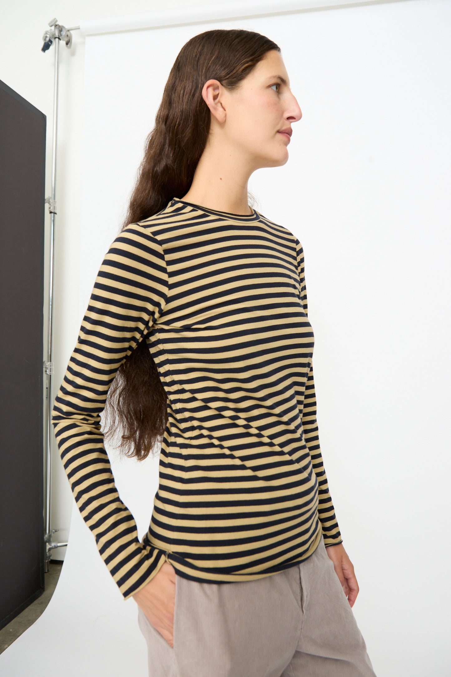 A person wearing the Labo.Art Striped Jersey Basic Longsleeve in Fennec, paired with light-colored pants, stands in profile against a white studio backdrop.