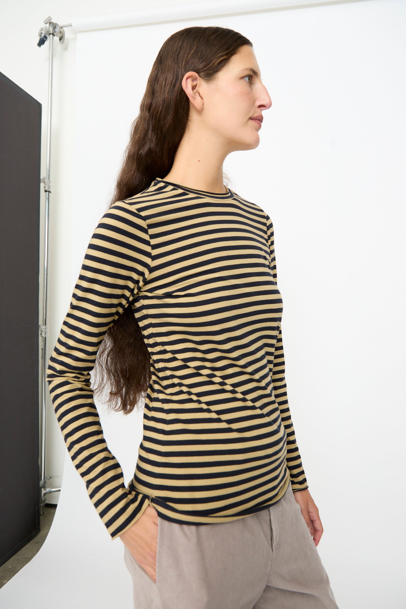 A person wearing the Labo.Art Striped Jersey Basic Longsleeve in Fennec, paired with light-colored pants, stands in profile against a white studio backdrop.