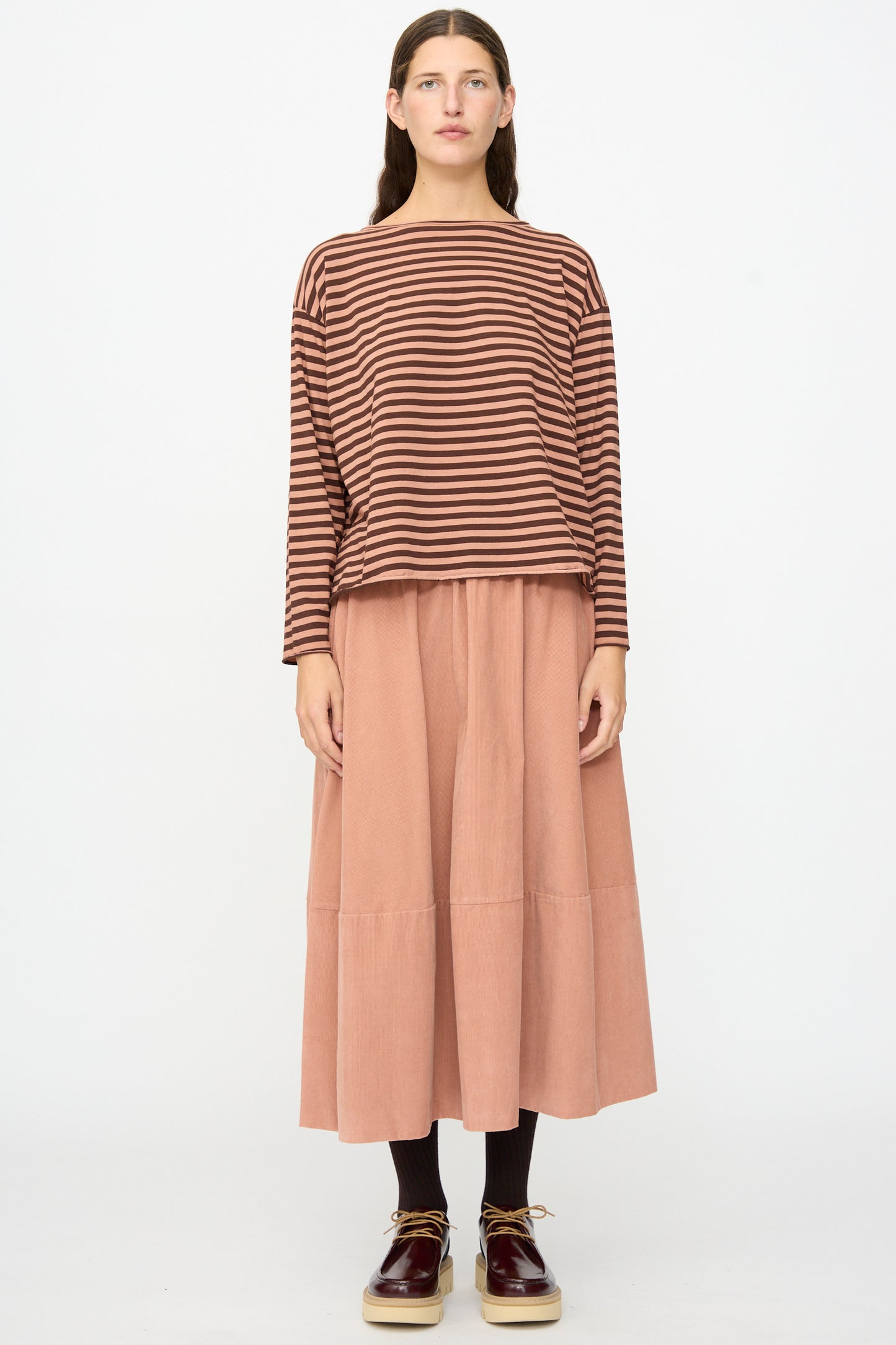 A woman stands facing forward against a plain white background, wearing the Striped Jersey Lola Sweater in Canyon by Labo.Art—a relaxed-fit, long sleeve brown and black striped top—paired with a peach-colored midi skirt, black socks, and burgundy shoes.