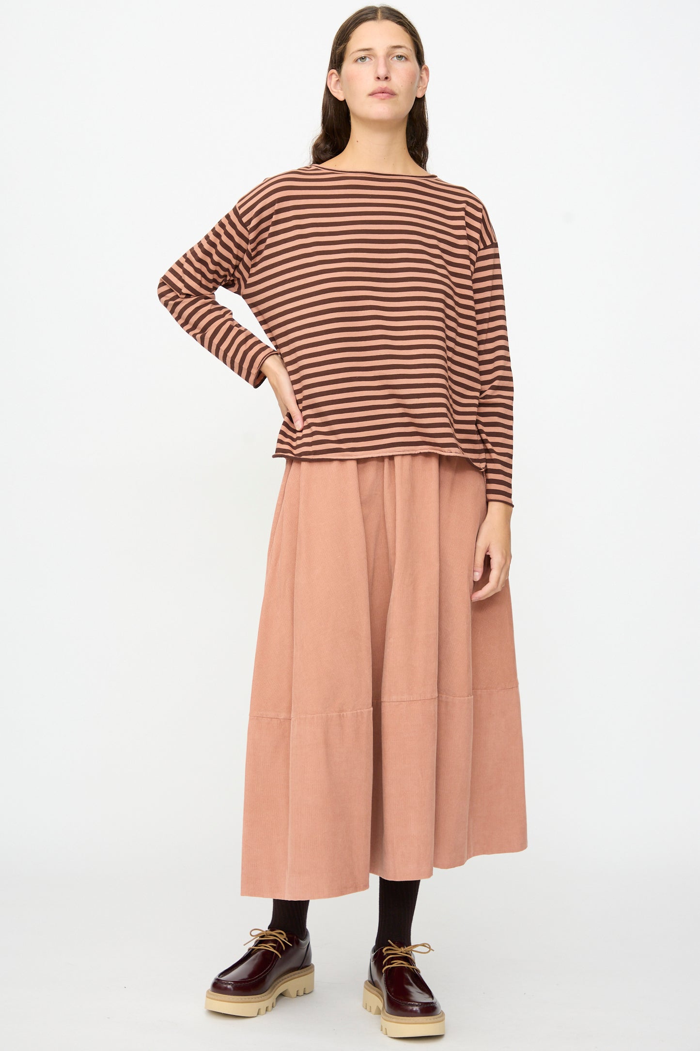 A person stands against a white background wearing the Striped Jersey Lola Sweater in Canyon by Labo.Art, paired with a peach-colored midi skirt, black tights, and brown platform shoes.