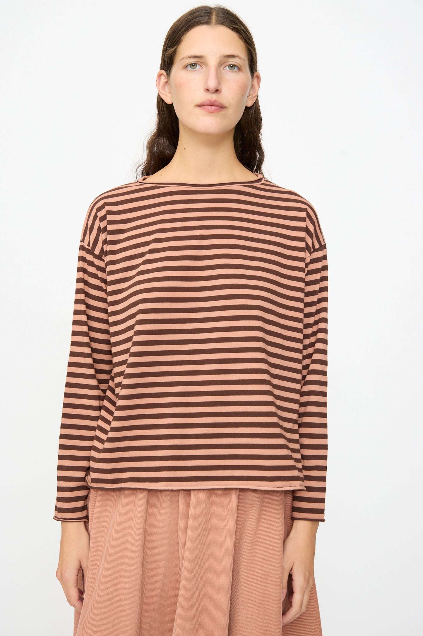 A model wears the Labo.Art Striped Jersey Lola Sweater in Canyon—a brown and pink striped long sleeve top with a round neckline—paired with a peach-colored skirt, standing against a plain white background.
