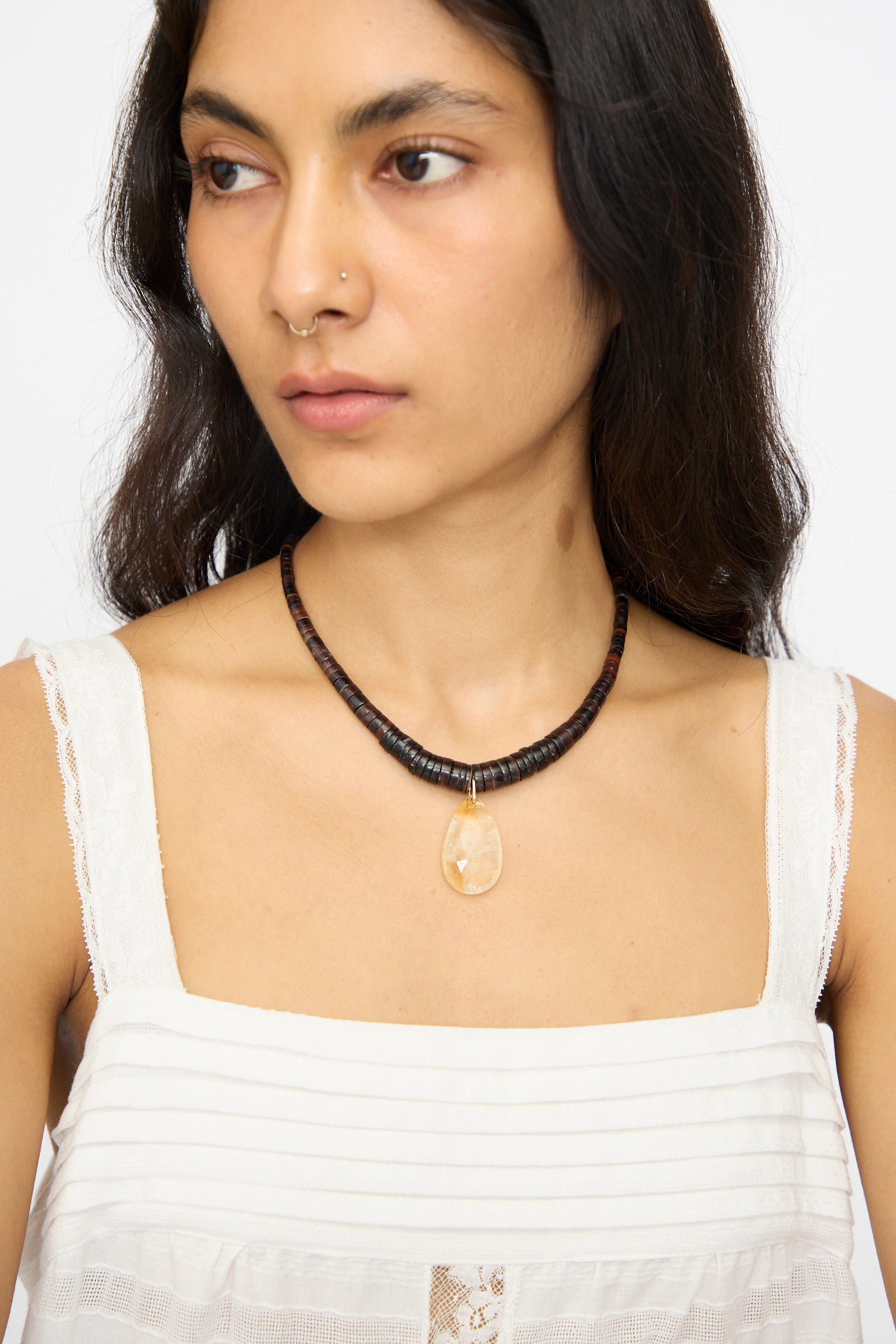 A woman in a white sleeveless top wears Laoli's The Drop Collar in Citrine, a handmade Parisian necklace featuring a dark beaded cord and large oval pendant, as she gazes slightly to the side.