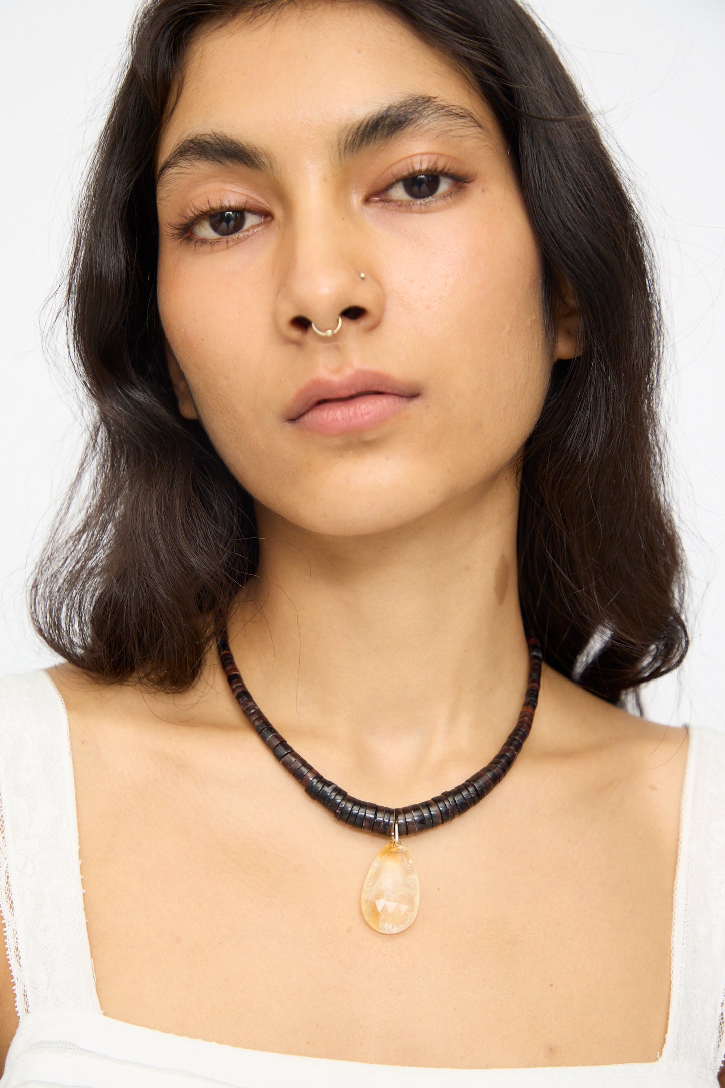 A woman with long dark hair and a neutral expression wears Laoli’s The Drop Collar in Citrine, a black beaded necklace with a citrine pendant, paired with a white top and a gold septum ring made from recycled gold jewelry.