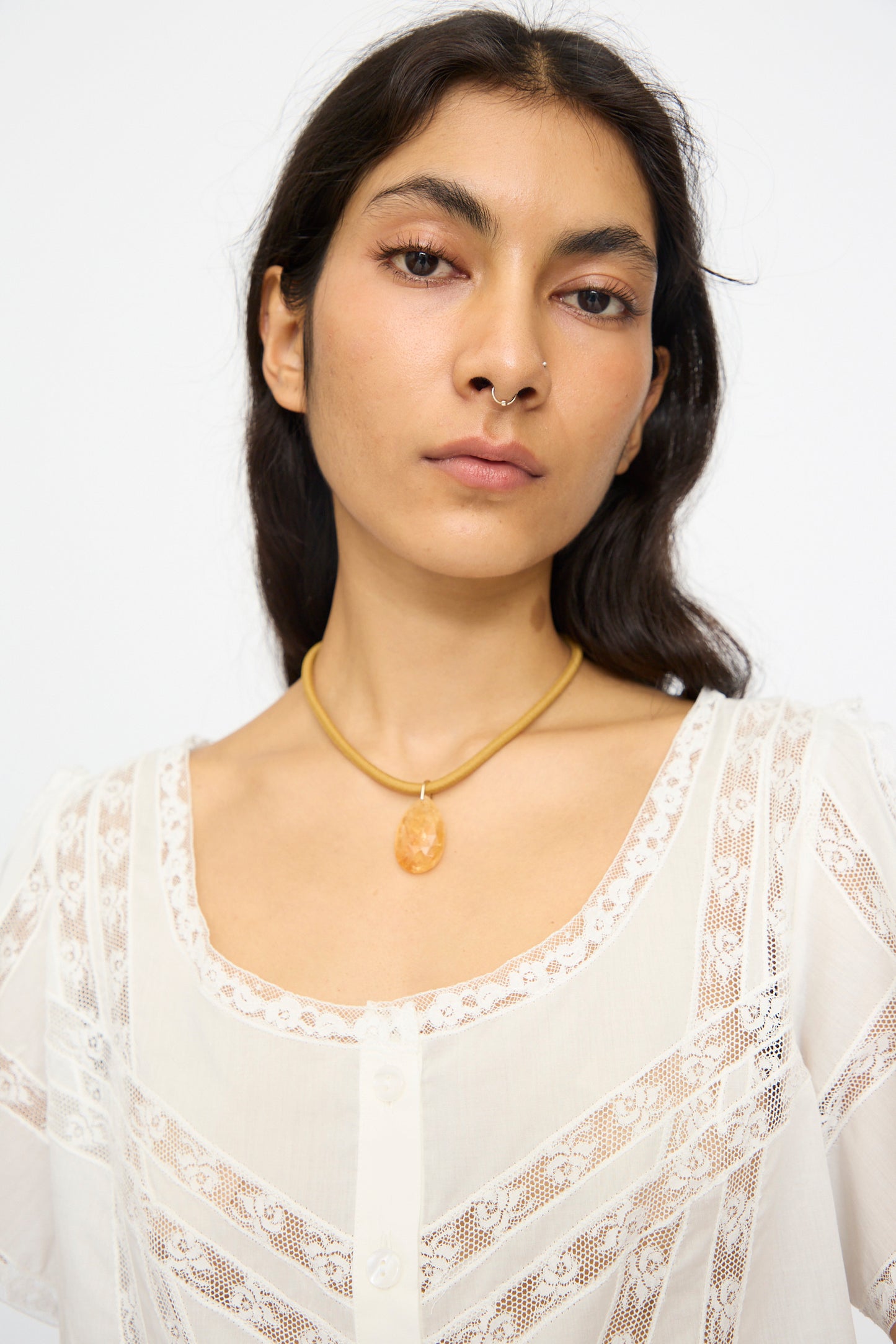 Woman with long dark hair wears Laoli's The Drop Cord in Tan and Citrine, an 18k gold choker with a peach-toned pendant, paired with a white lace blouse and nose ring against a plain light background.