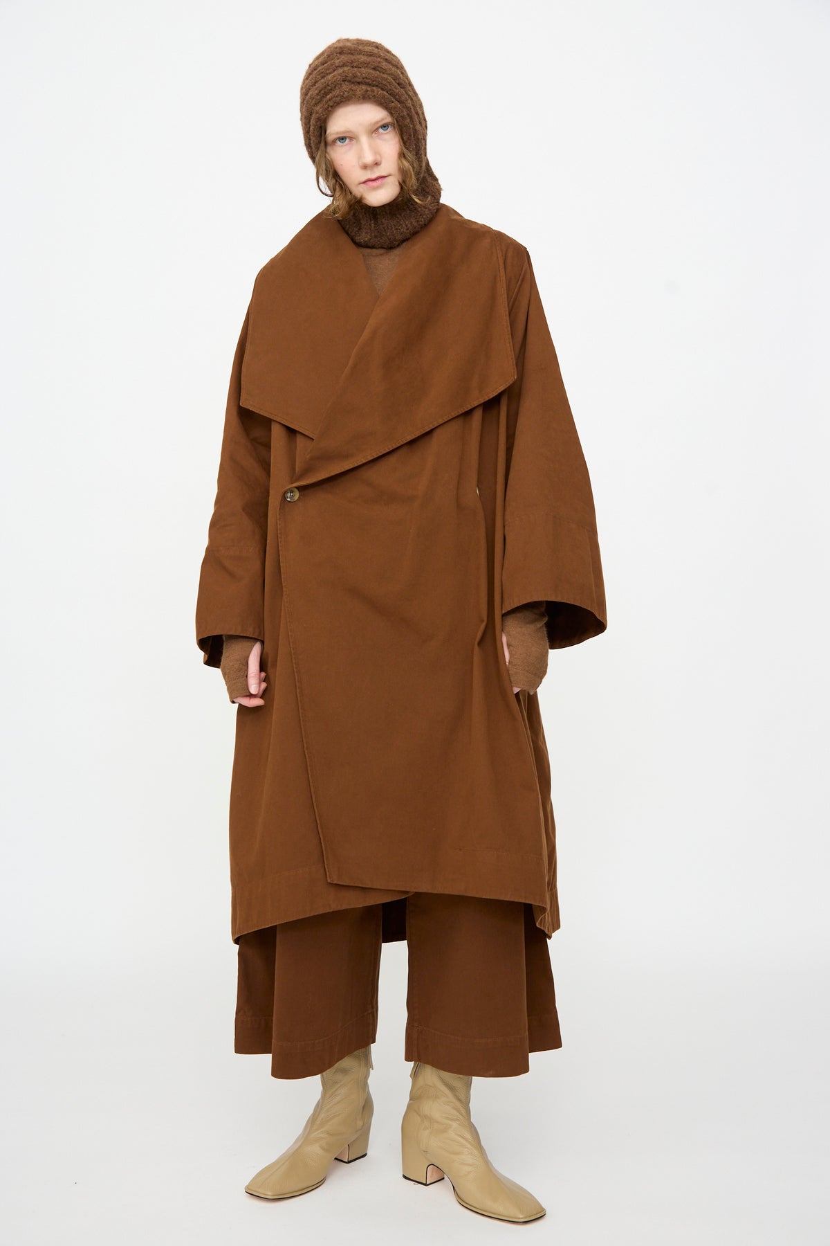 A person stands against a plain white background wearing a brown oversized coat, matching brown pants and hat, beige boots, and the Lauren Manoogian Handknit Rib Balaclava in Rust Melange.