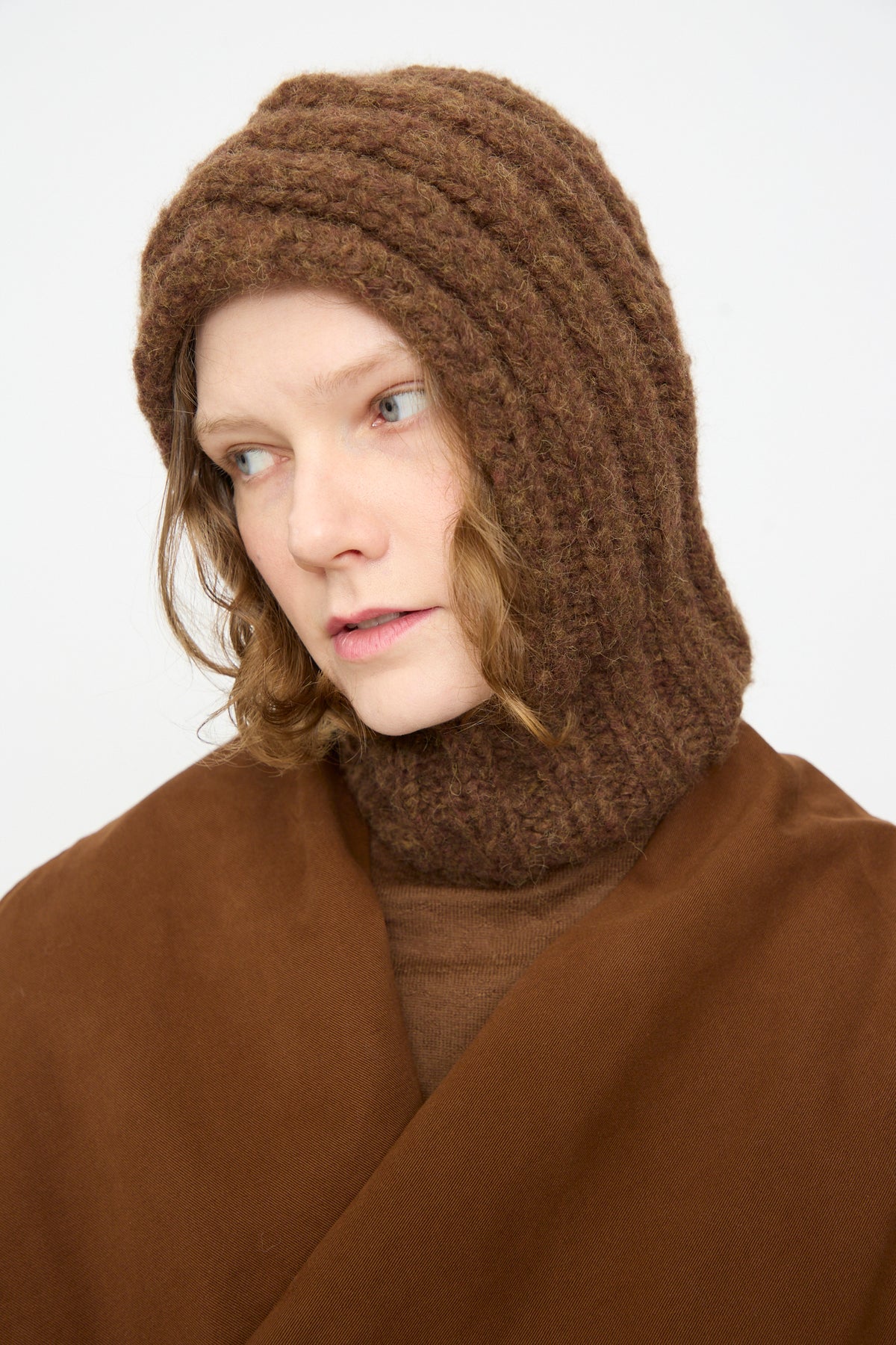 A person wearing the Lauren Manoogian Handknit Rib Balaclava in Rust Melange and layered brown clothing looks slightly to the side against a plain white background.