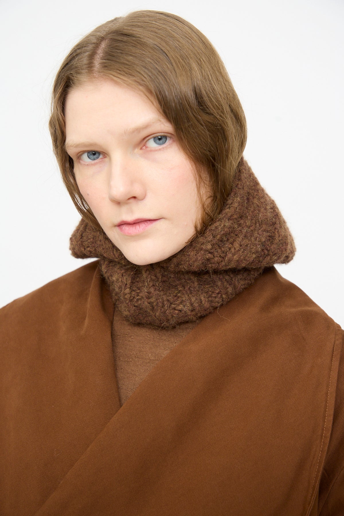 A person with short, light brown hair wears the Lauren Manoogian Handknit Rib Balaclava in Rust Melange and a brown coat, looking directly at the camera against a plain white background.