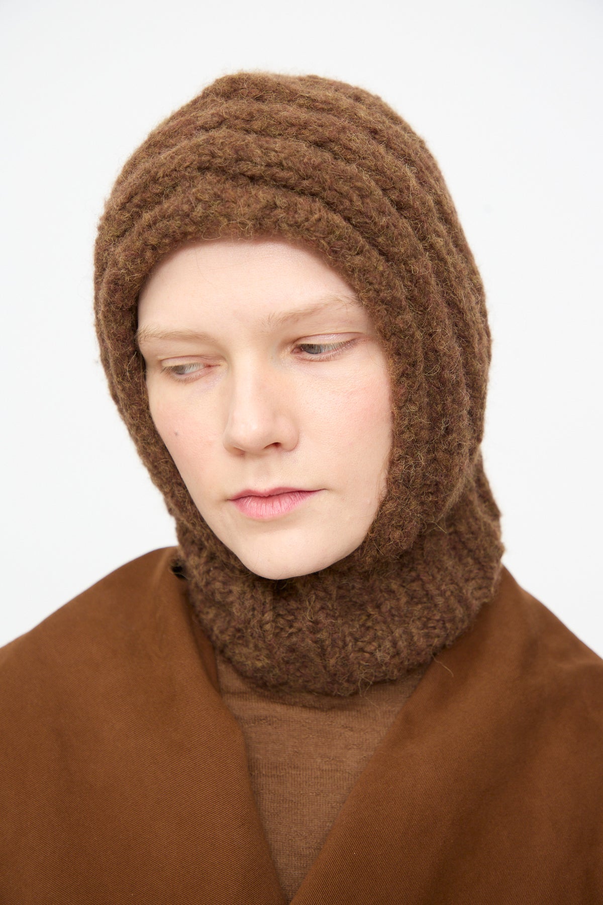 A person wears the Lauren Manoogian Handknit Rib Balaclava in Rust Melange—crafted from an alpaca blend—in rich brown tones, paired with matching brown clothing. They look slightly downward and to the side against a plain, light background. This distinctive accessory is made in Peru.