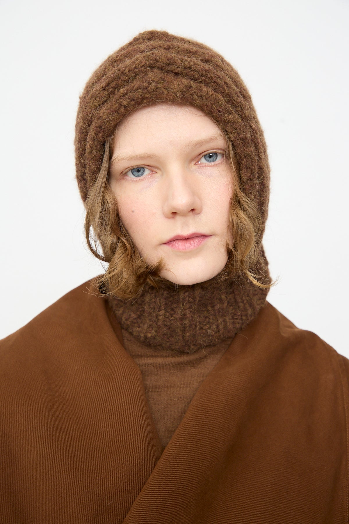 A person wearing the Lauren Manoogian Handknit Rib Balaclava in Rust Melange and coordinating brown attire, gazes directly at the camera against a simple light backdrop.
