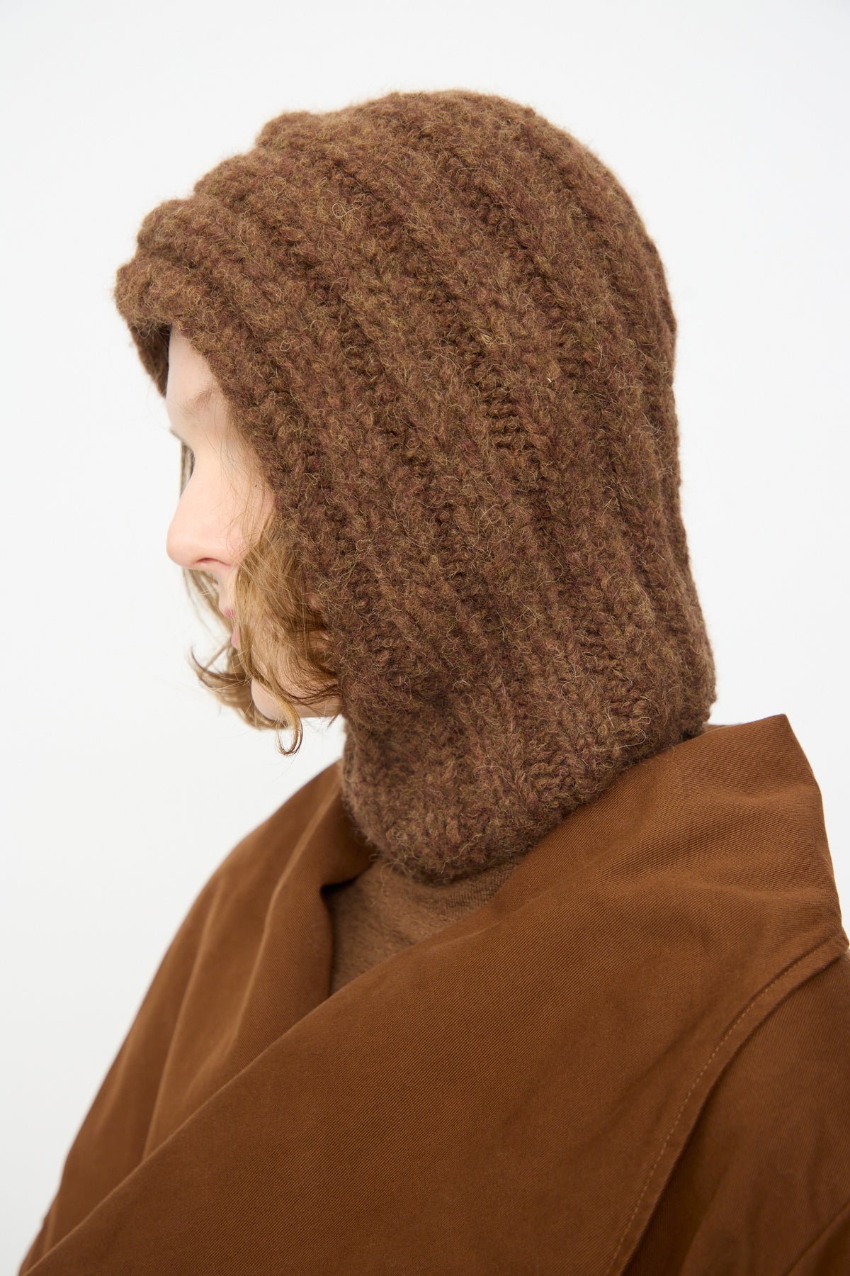A person in profile wears the Lauren Manoogian Handknit Rib Balaclava in Rust Melange and a brown coat, with light brown hair visible against a neutral background.