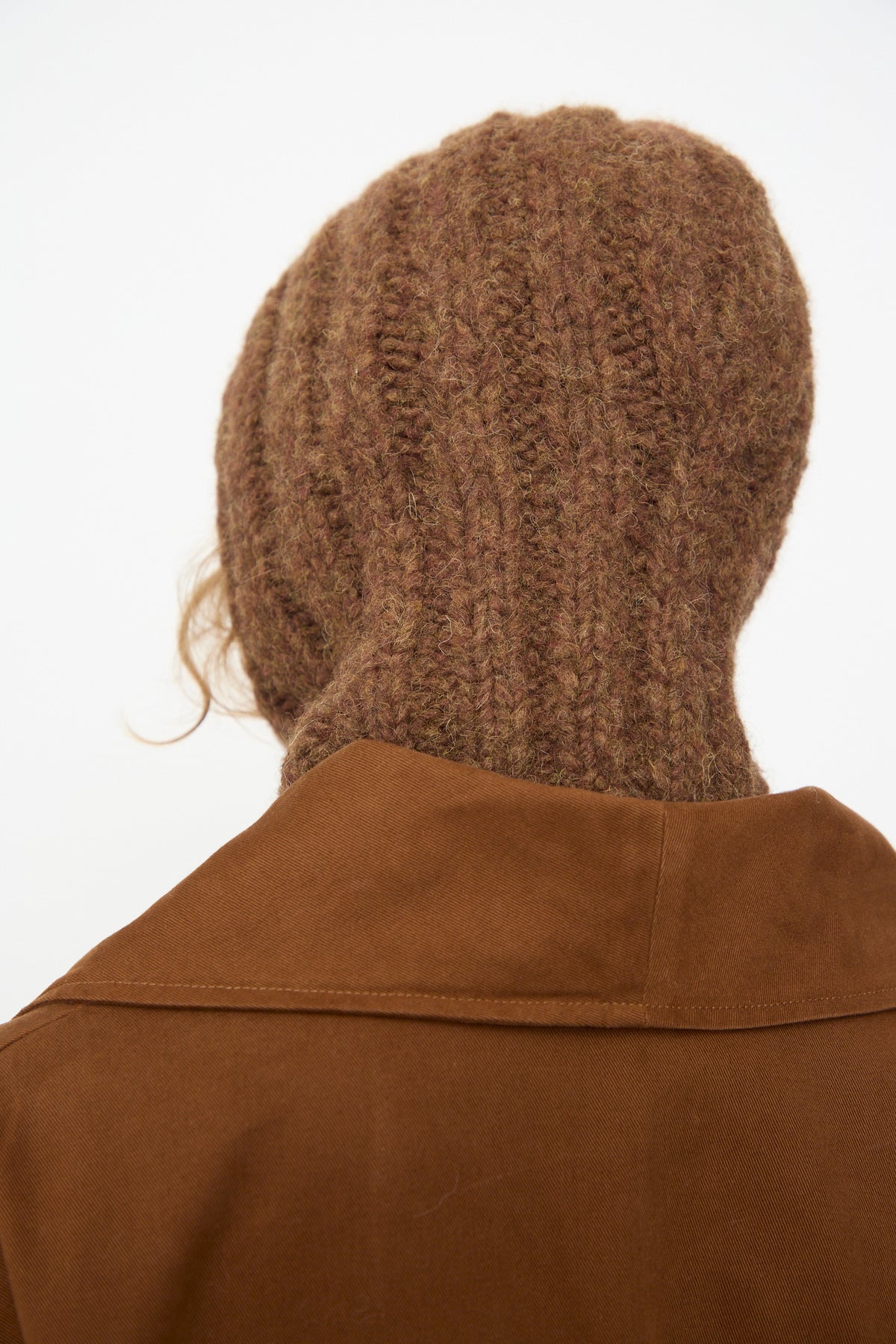 A person wears the Lauren Manoogian Handknit Rib Balaclava in Rust Melange with a brown coat, photographed from behind against a plain background. Crafted in Peru from a soft alpaca blend, the cozy balaclava adds distinctive texture to the look.